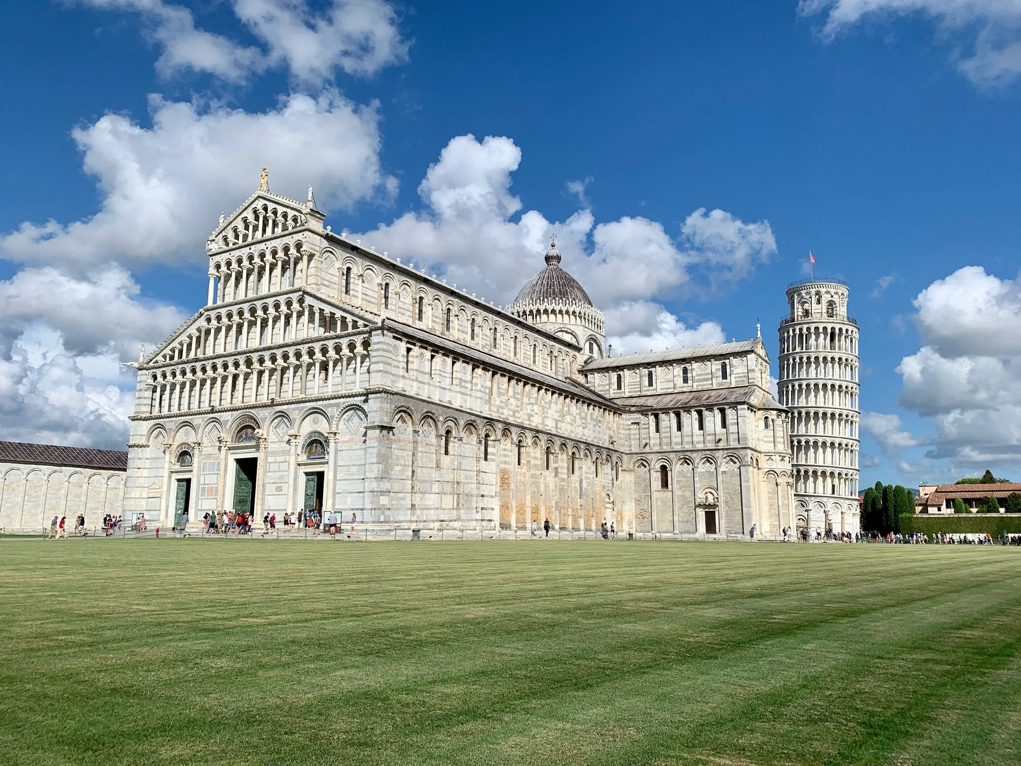 The Leaning Tower of Pisa, Pisa Cathedral, and Piazza dei Miracoli with people walking around and a partly cloudy sky. Tuscany, Italy.