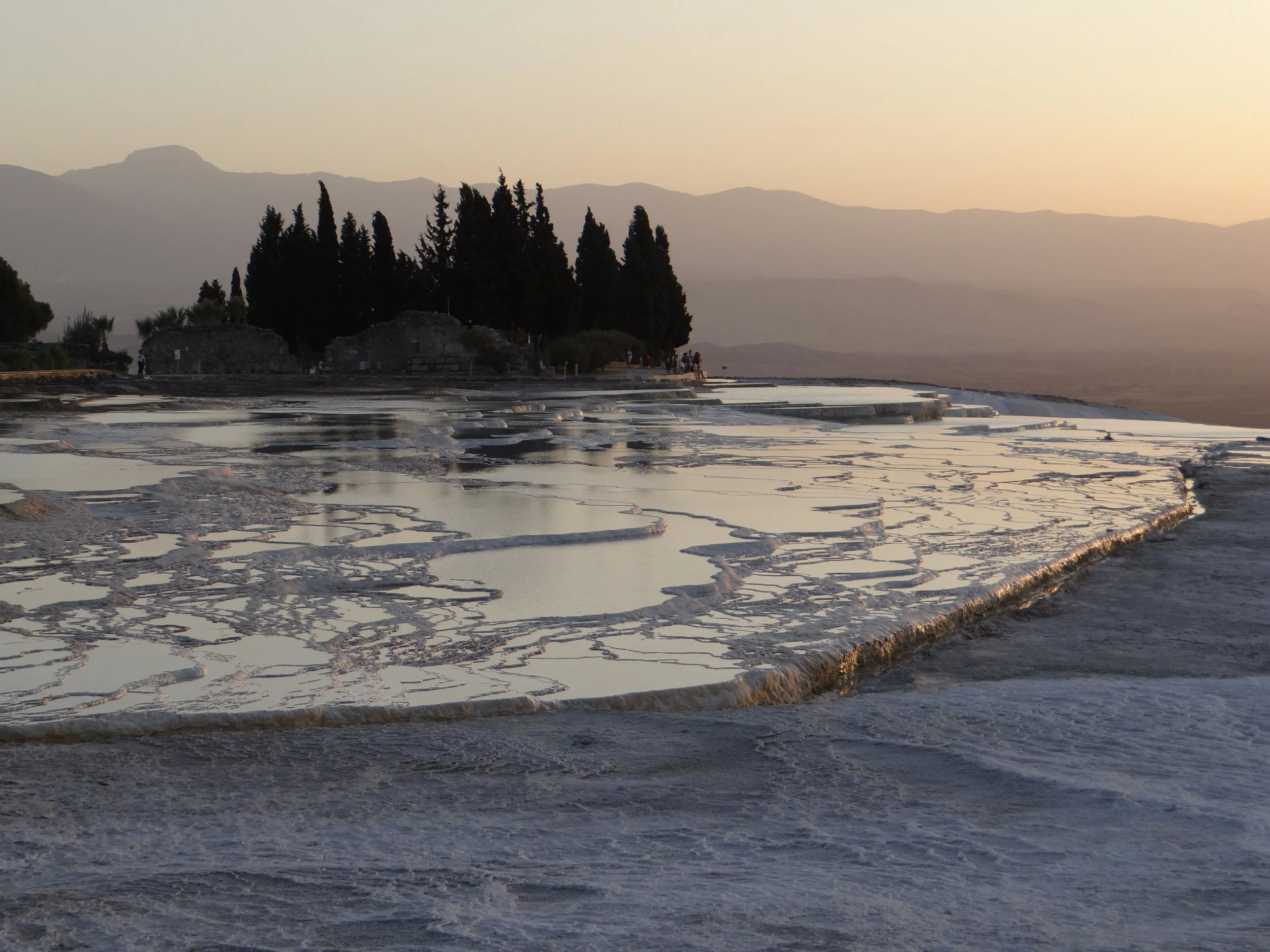 Salt terraces at Pamukkale, Turkey, during sunset, with trees and mountains in the background.