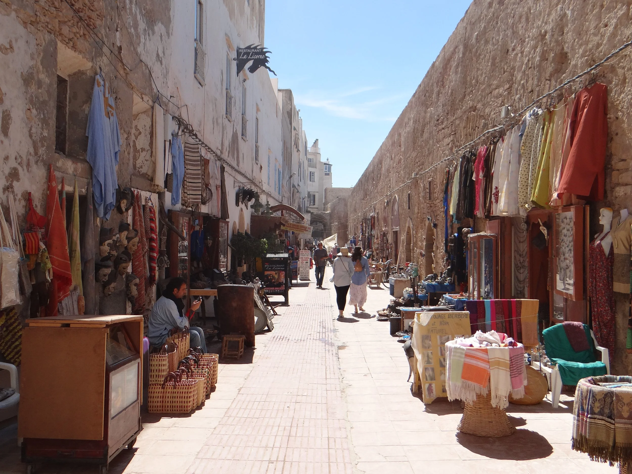 Street market with clothing and crafts displayed outside shops along a narrow alley in the historic area of Essaouira, Morocco, with tourists walking and browsing.