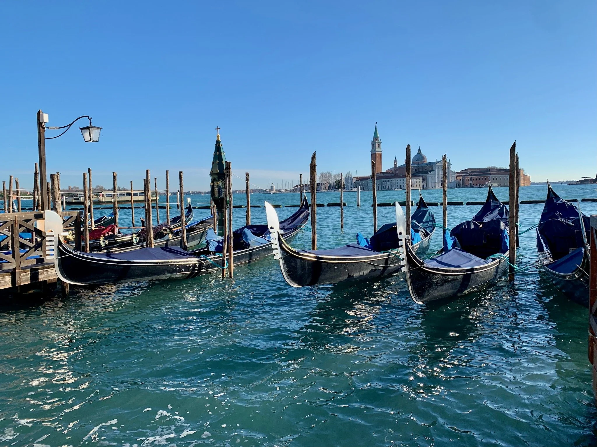 Gondolas moored at a wooden dock on a sunny day in Venice, Italy, with historic buildings and a church steeple visible across the water in the background.