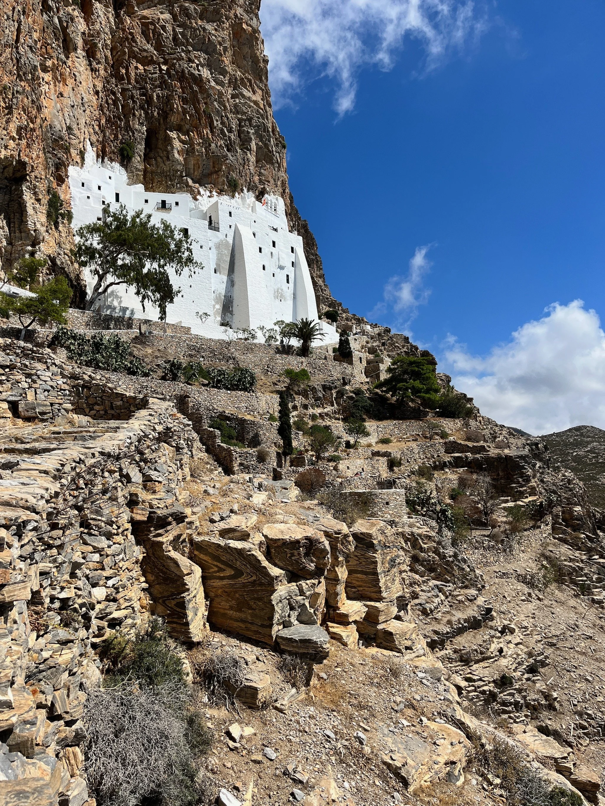 The white monastery of Hozoviotissa on Amorgos island, in the Cyclades, Greece, built into a rocky cliffside, with terraced stone pathways and sparse vegetation, under a partly cloudy blue sky.