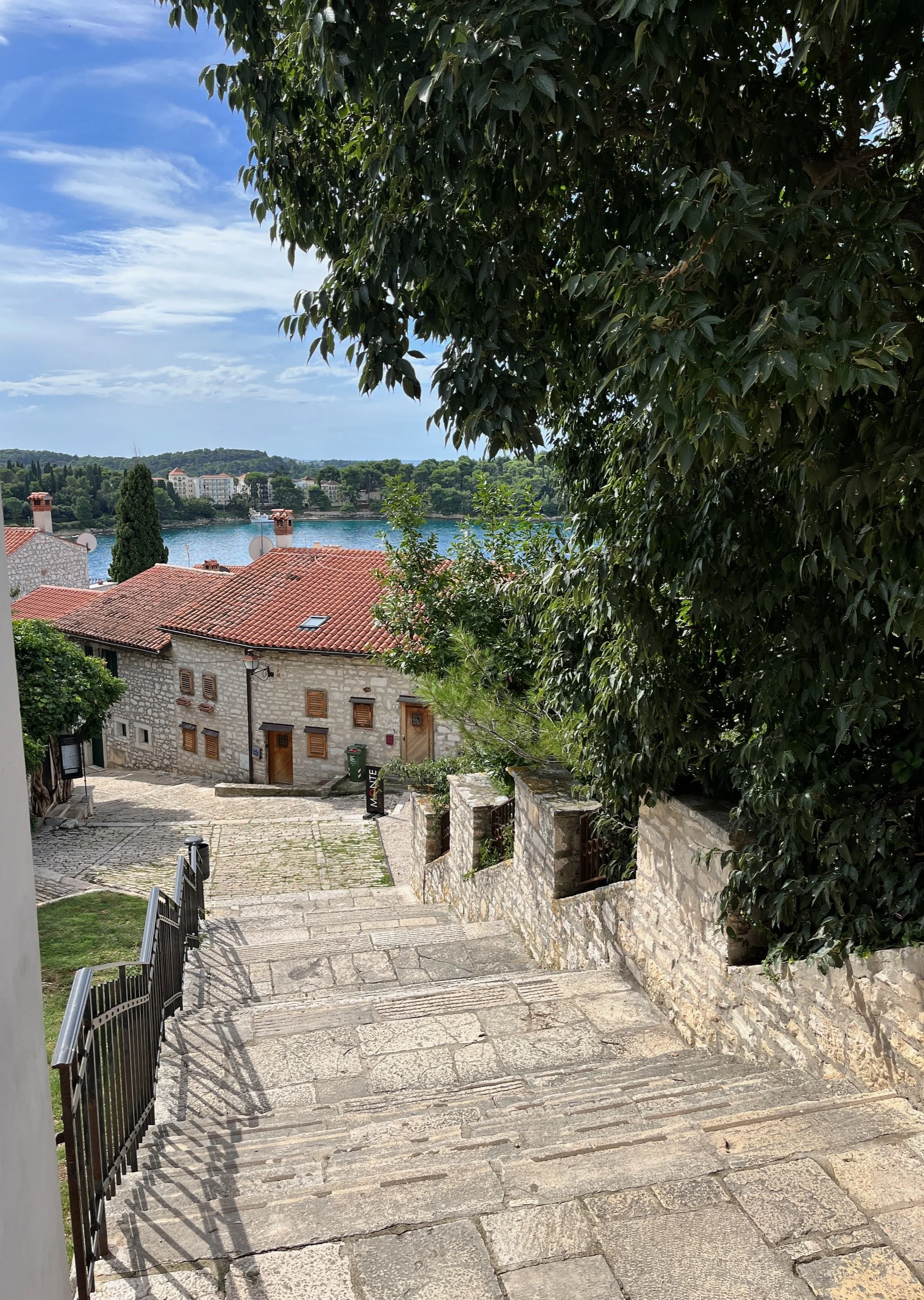 Stone stairway leading down to a square with a stone building with red-tiled roof, in Rovinj, Croatia, overlooking a body of water with trees and buildings in the background, partially shaded by large green trees.