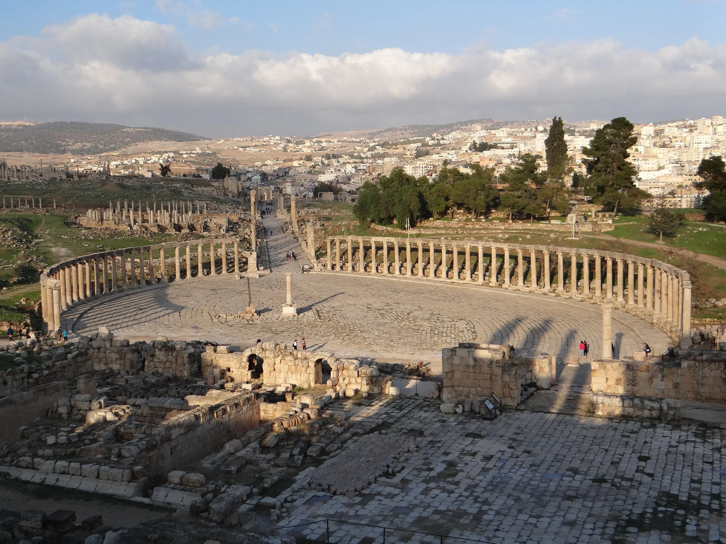 Ancient ruins of a circular stone Roman square with columns, at the archeological site of Jerash, Jordan, surrounded by trees and cityscape in the background under partly cloudy sky.