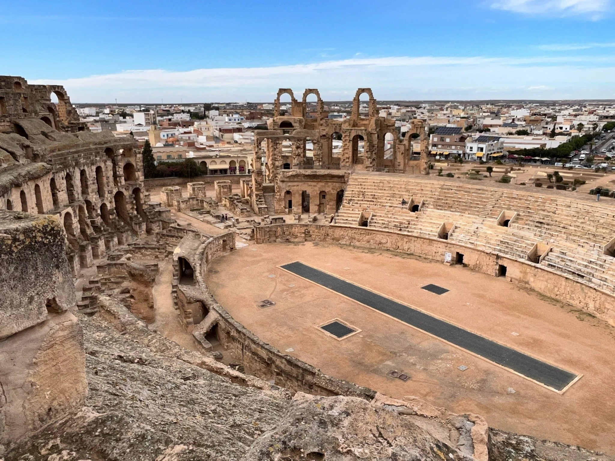 The ancient Roman amphitheater of El Jem, Tunisia, with ruins with stone seating, arches, and a central stage, set against the modern town of El Jem with white buildings under a blue sky.