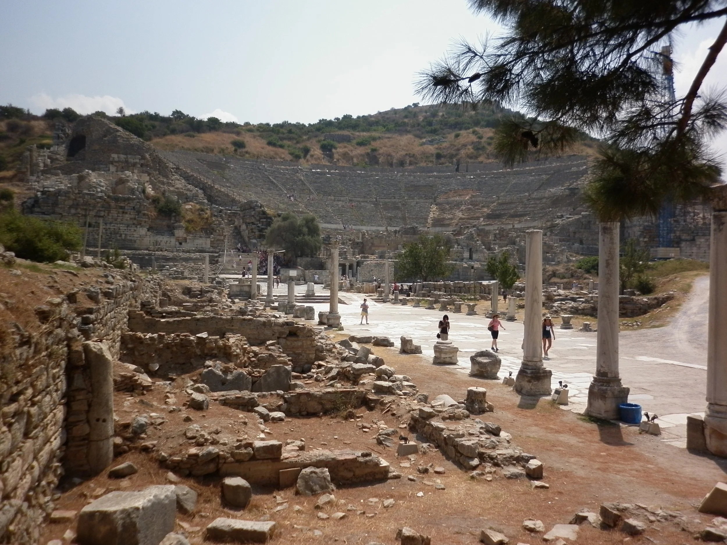 Ancient ruins of a Roman theater and street in Hierapolis, Turkey, with stone columns, some standing and some fallen, under a clear sky with a hillside in the background. There are tourists exploring the site.