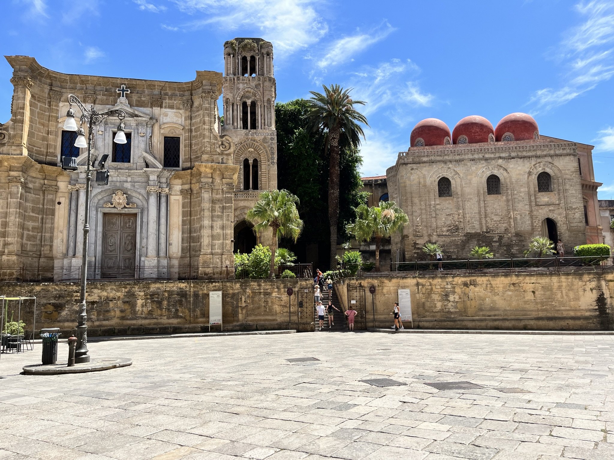 The Church of La Martorana and the Church of San Cataldo  in Palermo, Sicily in Italy, with a bell tower, three red domes, palm trees, and a blue sky with clouds, people walking in front on a paved square.