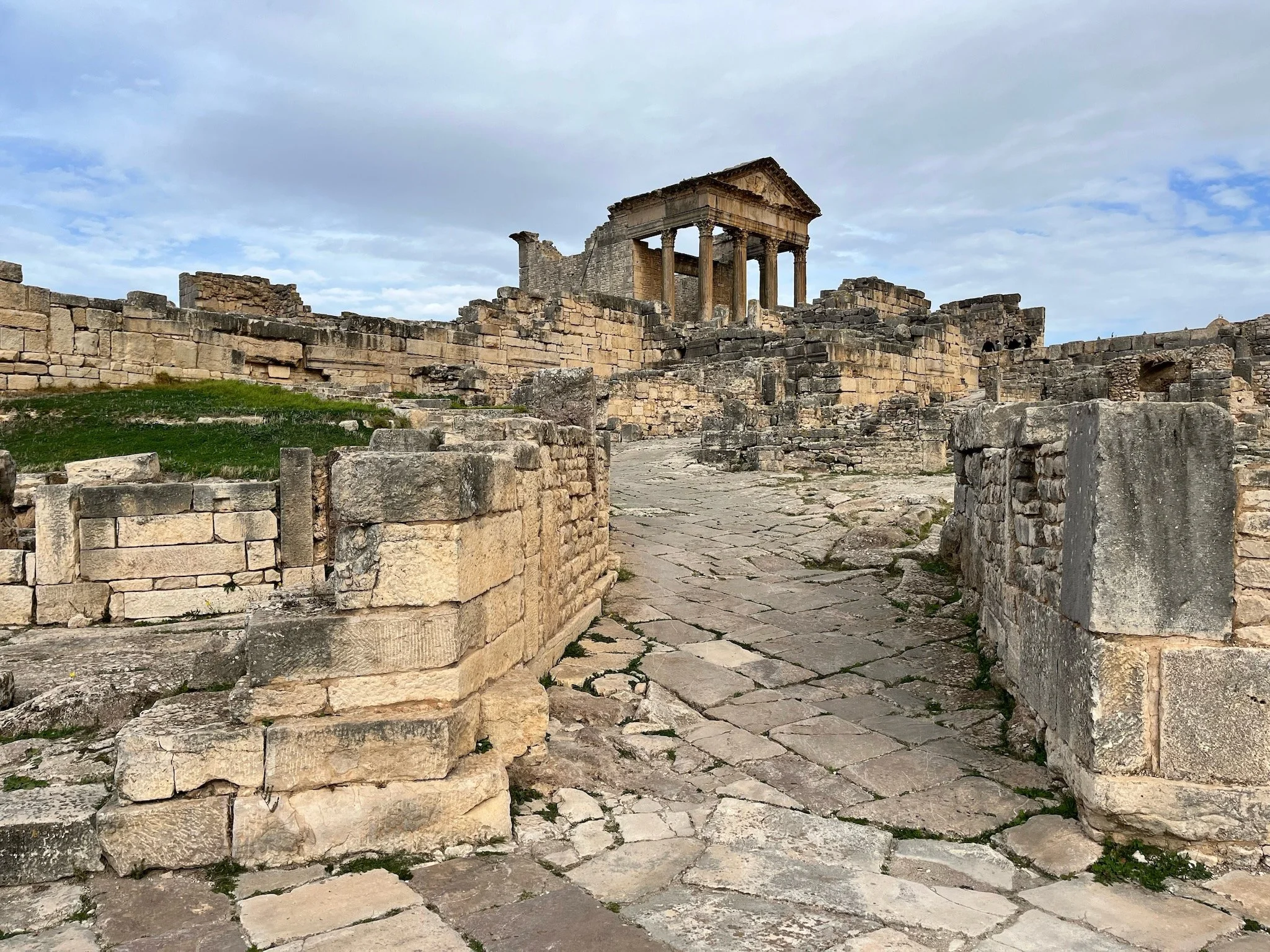 Ancient ruins with stone steps leading to a Roman-style temple with columns, in Dougga, Tunisia, under a cloudy sky.
