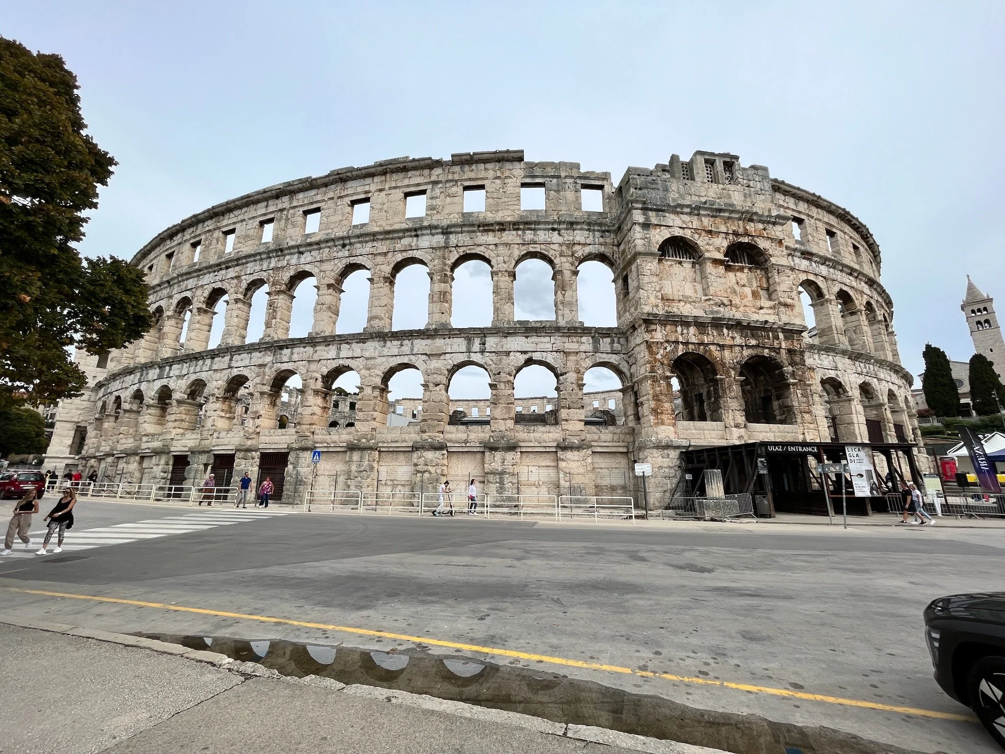 The ancient Roman amphitheater of the city of Pula, in Istria, Croatia, with some people walking nearby and a small puddle reflecting part of the structure on the street in front.