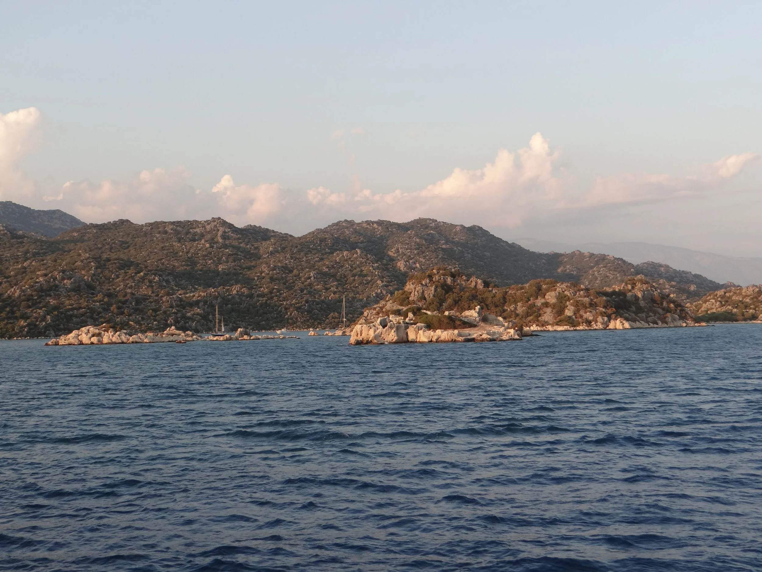 A coastal scene with rocky hills and mountains in the background, and the Mediterranean Sea in the Turkish Riviera, between Bodrum and Antalya, Turkey. In the foreground there are small waves and a few boats anchored near the shoreline.