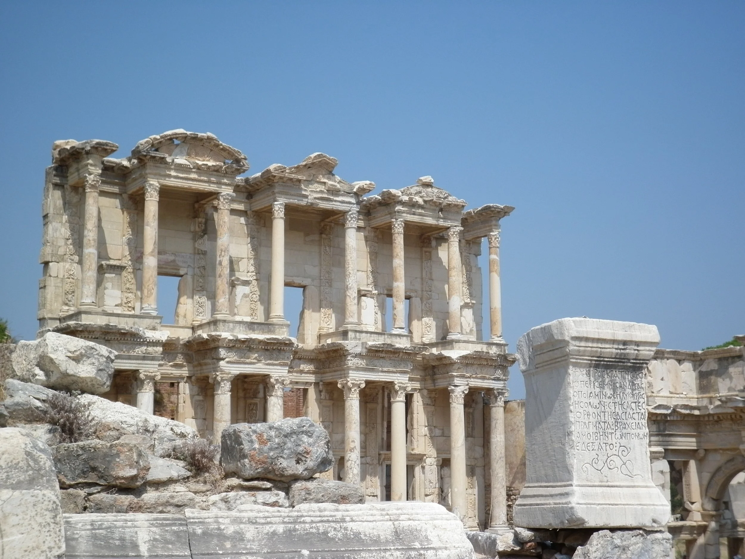 The ancient ruins of the library of Celsus, in Ephesus, Turkey, a stone building with tall columns, decorative carvings, and an inscribed stone slab, under a clear blue sky.