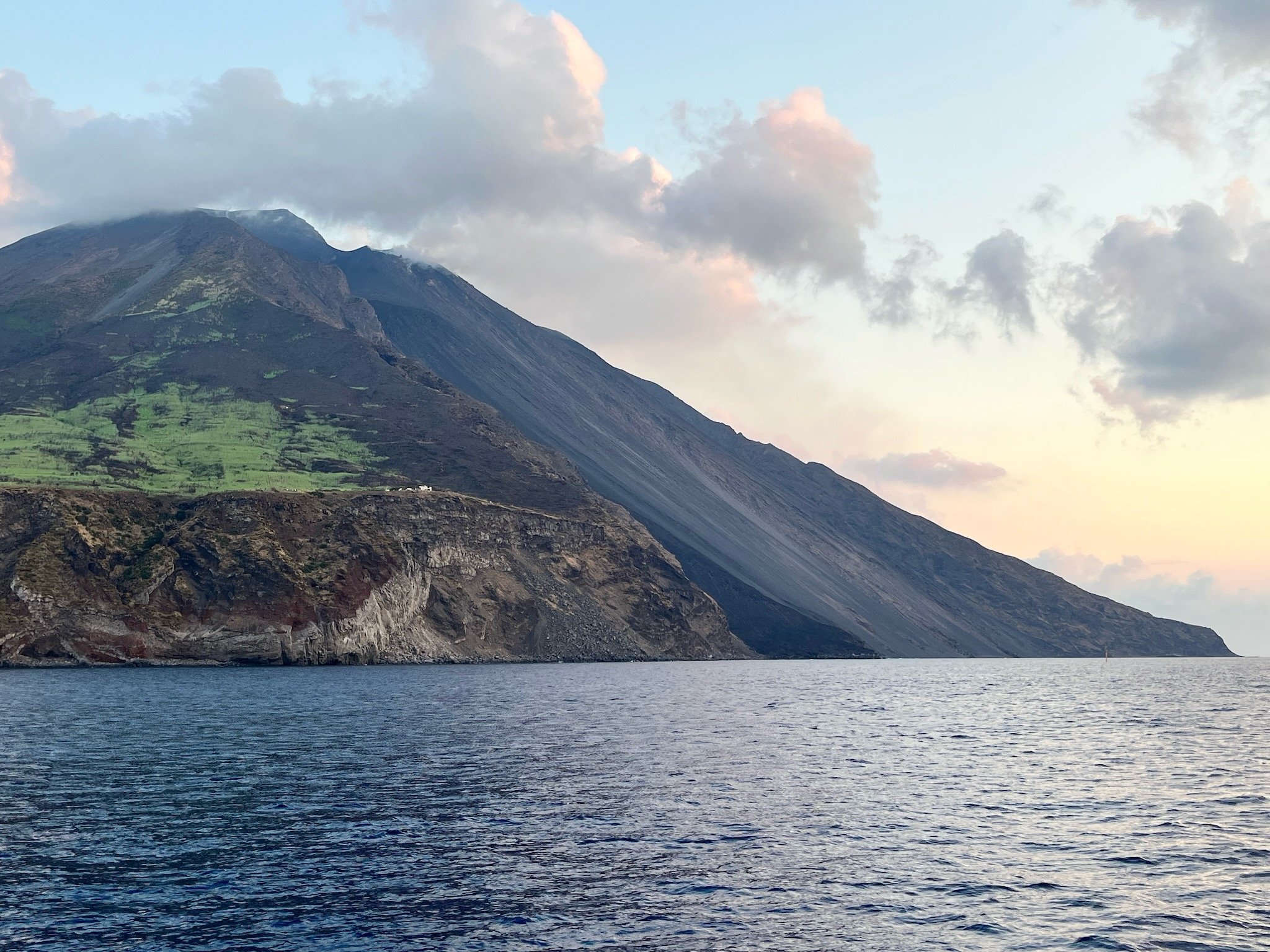 Scenic view over la sciara del fuoco and the volcano on Stromboli island, in Sicily in Italy in the Aeolian islands with patches of green vegetation, a partly cloudy sky, and the Mediterranean Sea in the foreground.