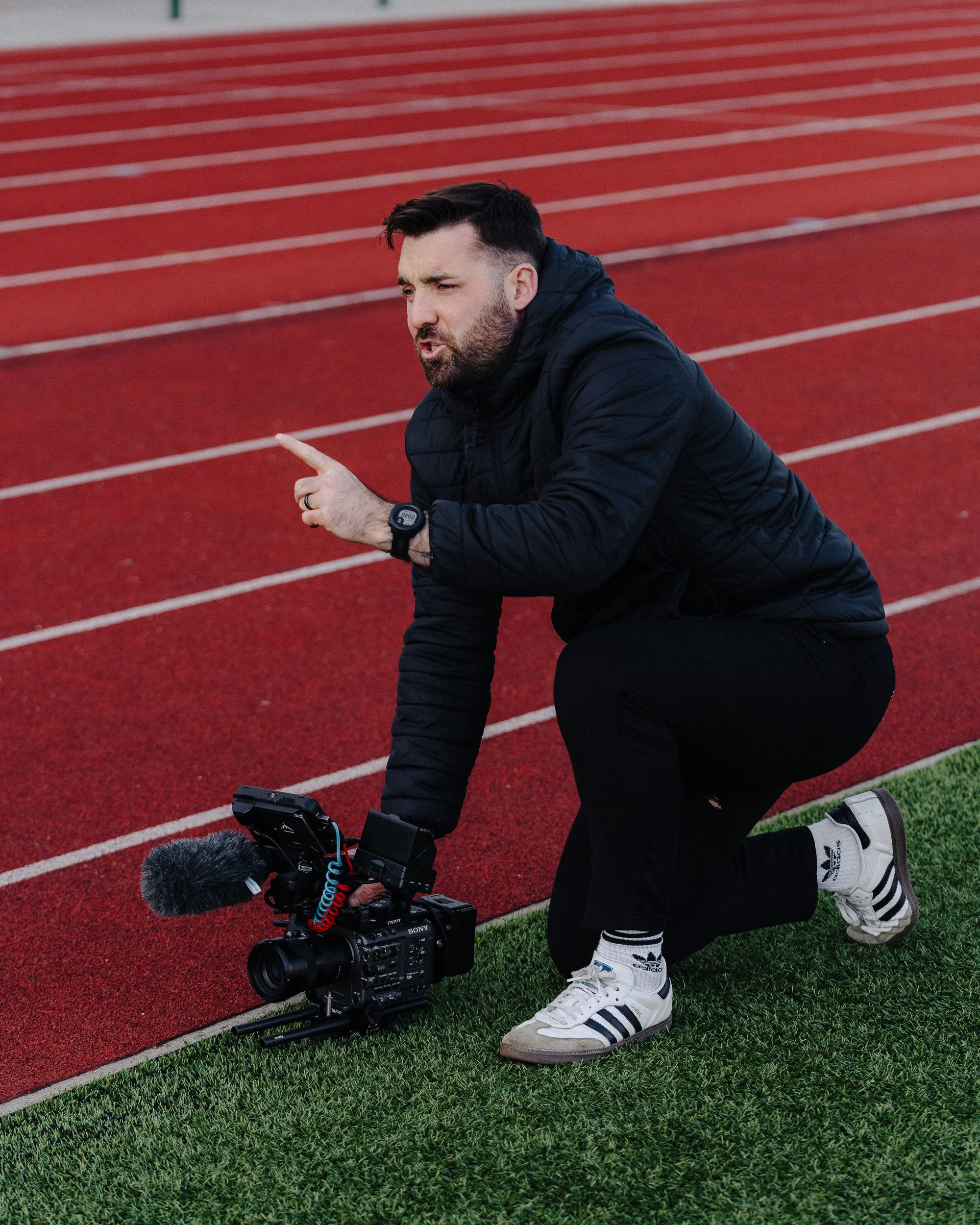Man kneeling on a running track with video camera, pointing and speaking, wearing black athletic clothes and Adidas sneakers.