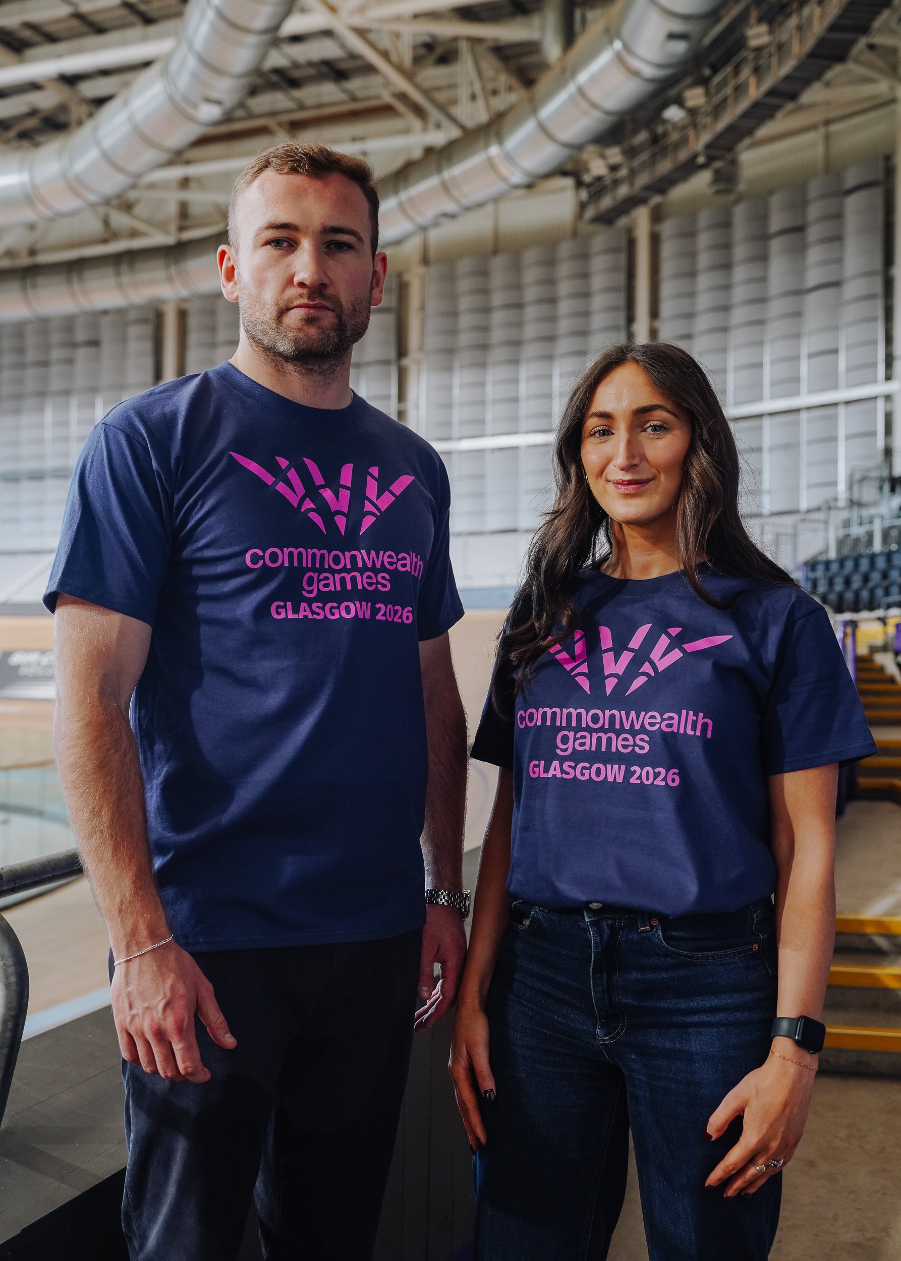 Two people wearing navy blue T-shirts with pink text that reads 'commonwealth games GLASGOW 2026' standing inside a large indoor sports arena with ventilation ducts visible in the background.