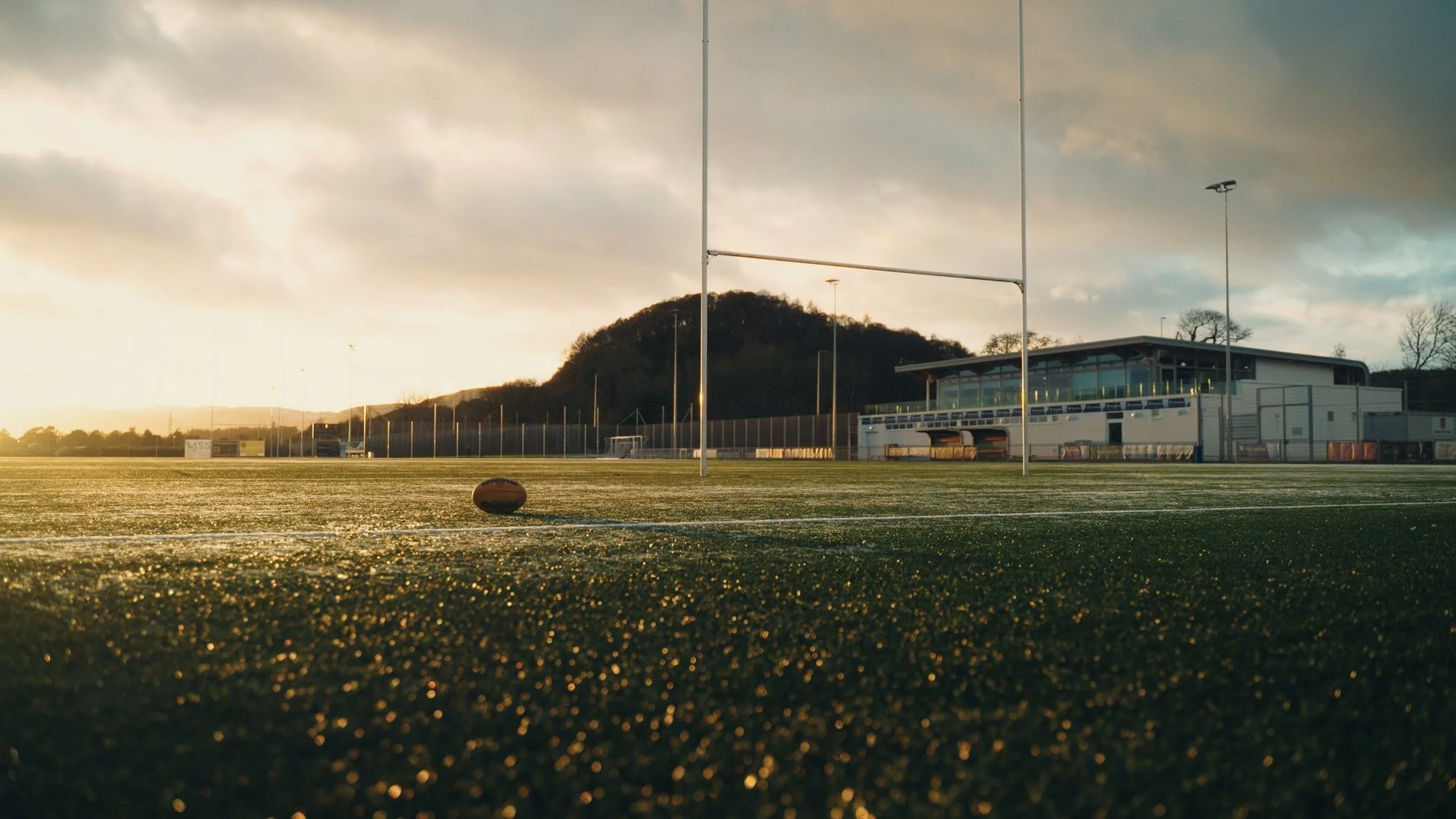 Empty rugby field at sunset with rugby ball on the ground and goalposts in the foreground. Spectator stands and a building are in the background, with a hill and cloudy sky overhead.
