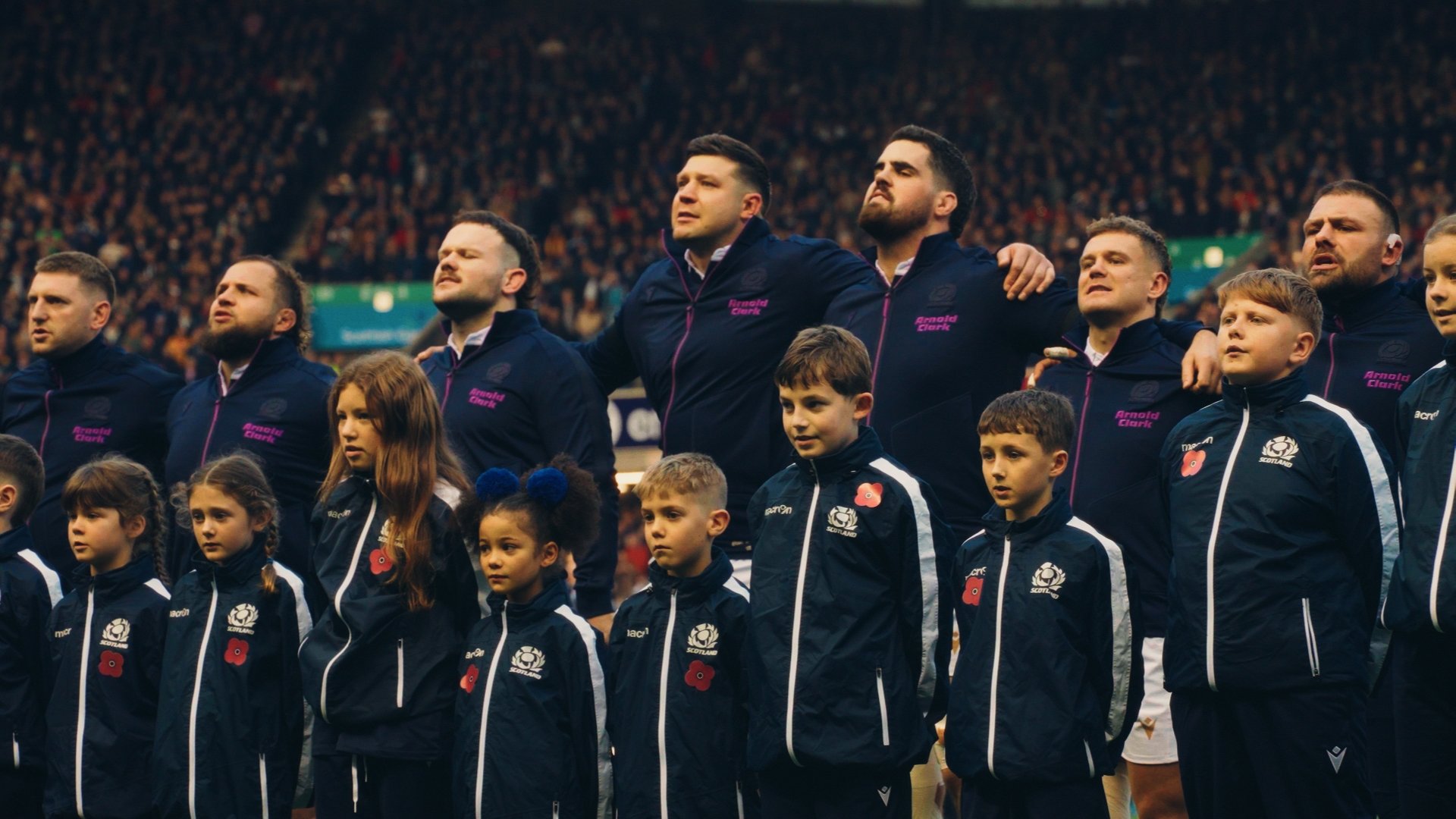 A group of rugby players and children standing with arms around each other's shoulders during a national anthem at a sports arena filled with spectators.
