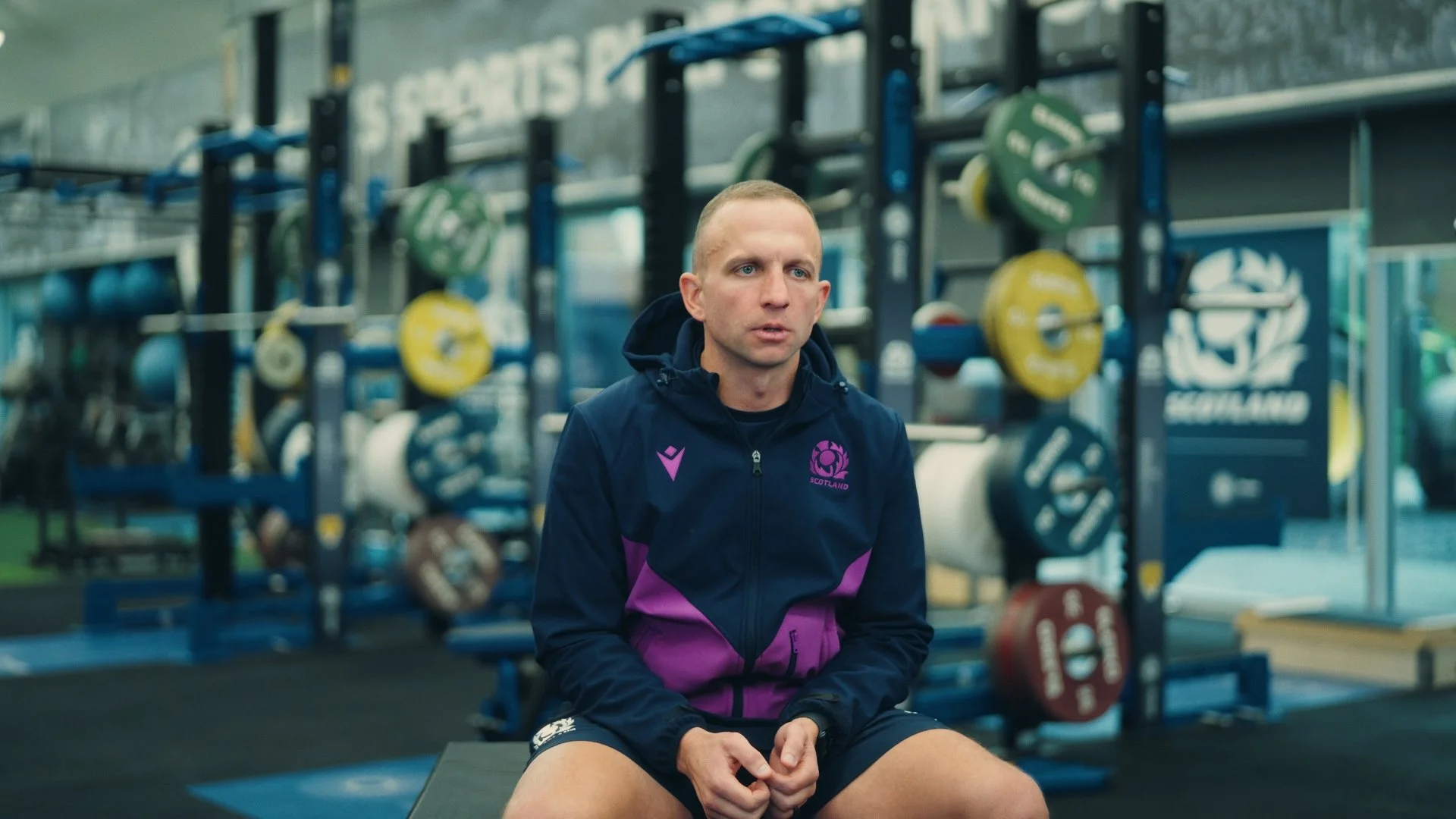 A man wearing a navy blue and purple Scotland sports jacket sitting on a bench in a gym.