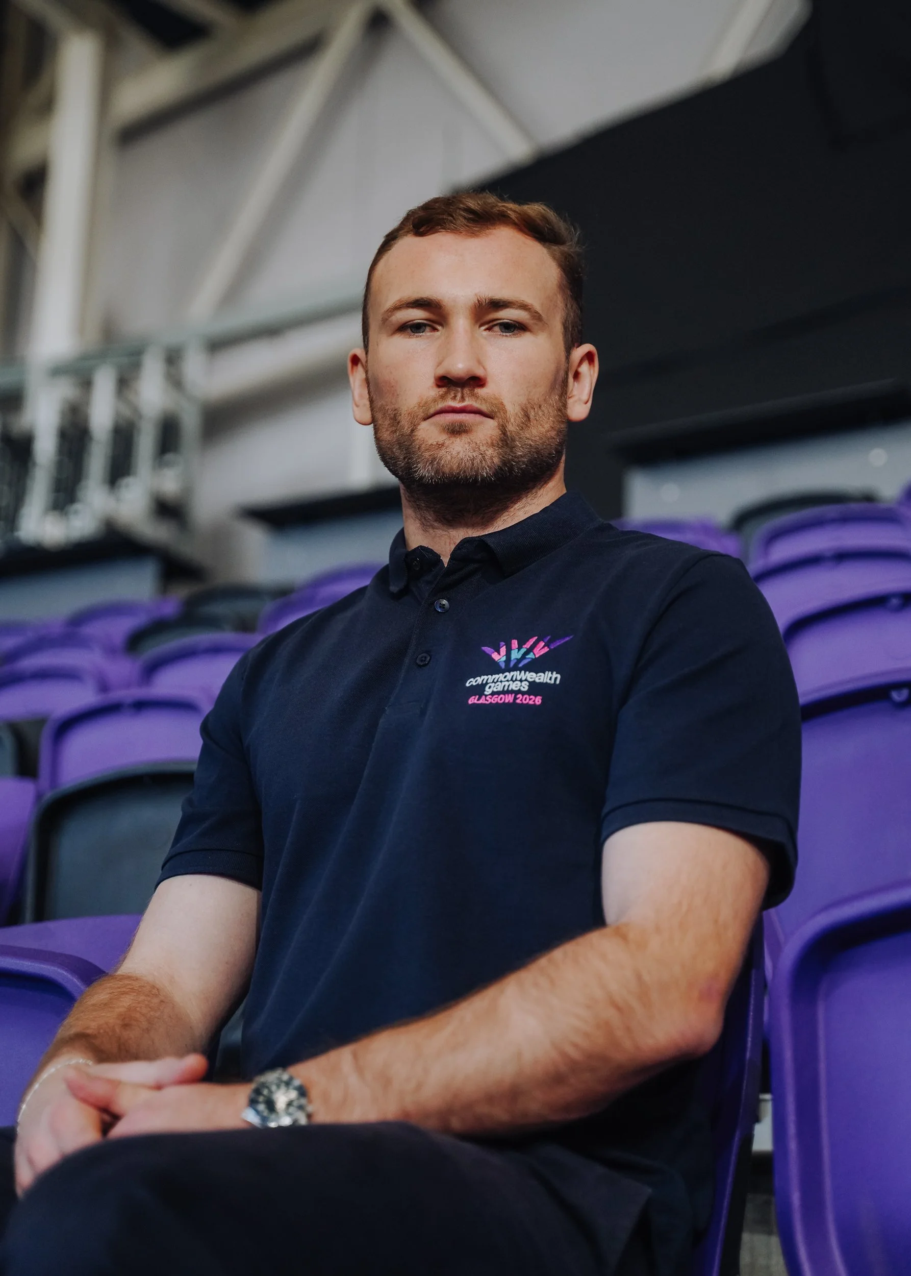 A man with short brown hair and a beard sitting on purple stadium seats inside an arena. He is wearing a dark navy polo shirt with a logo that says 'Commonwealth Games Glasgow 2026' on it.