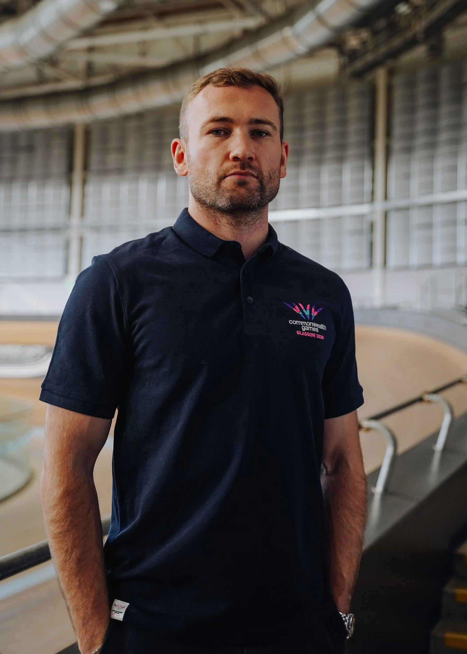 A man in a black polo shirt standing indoors, with a serious expression, in a sports arena with a curved roof and railing in the background.
