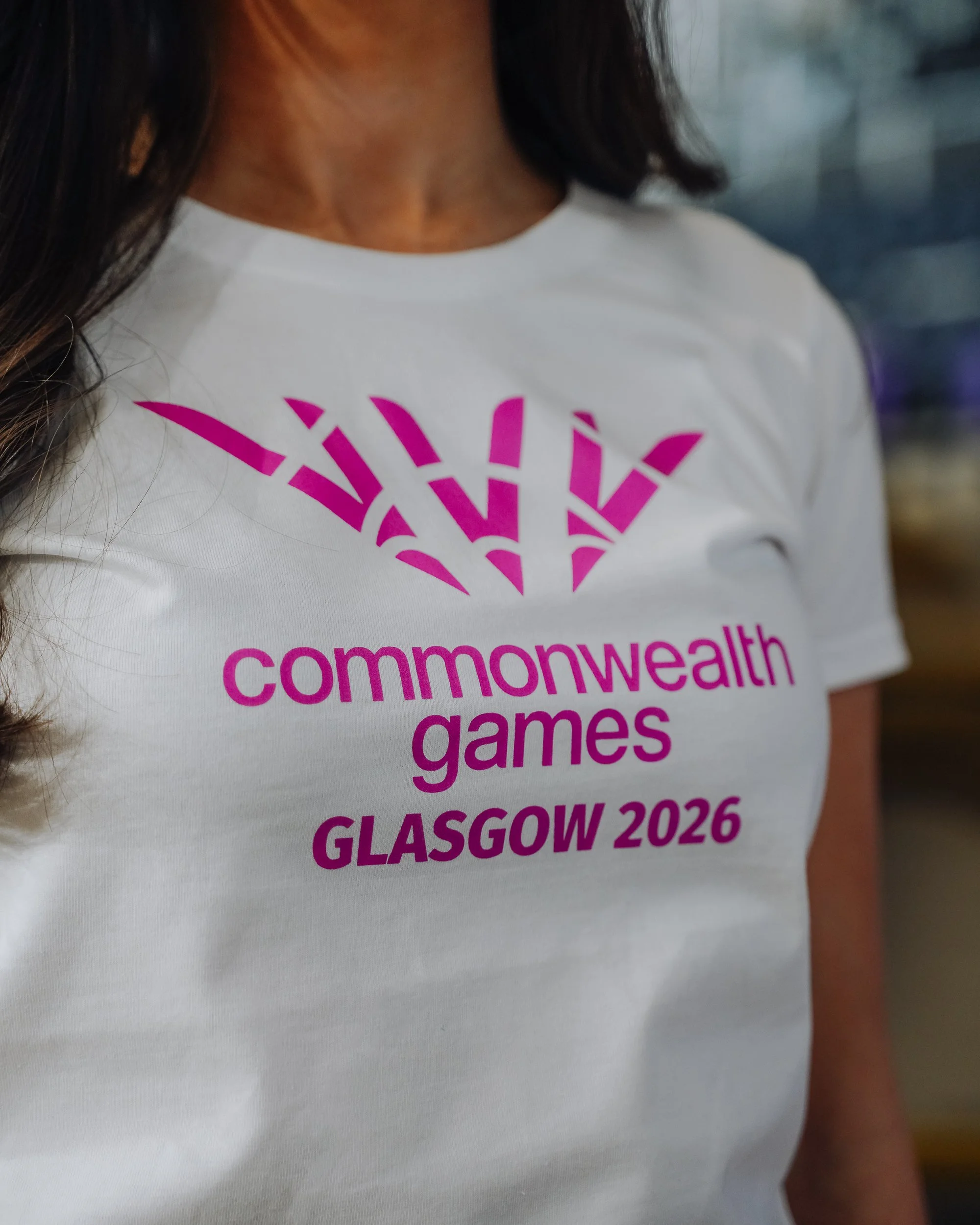 Close-up of a white T-shirt with pink text and logo, reading 'Commonwealth Games Glasgow 2026'.