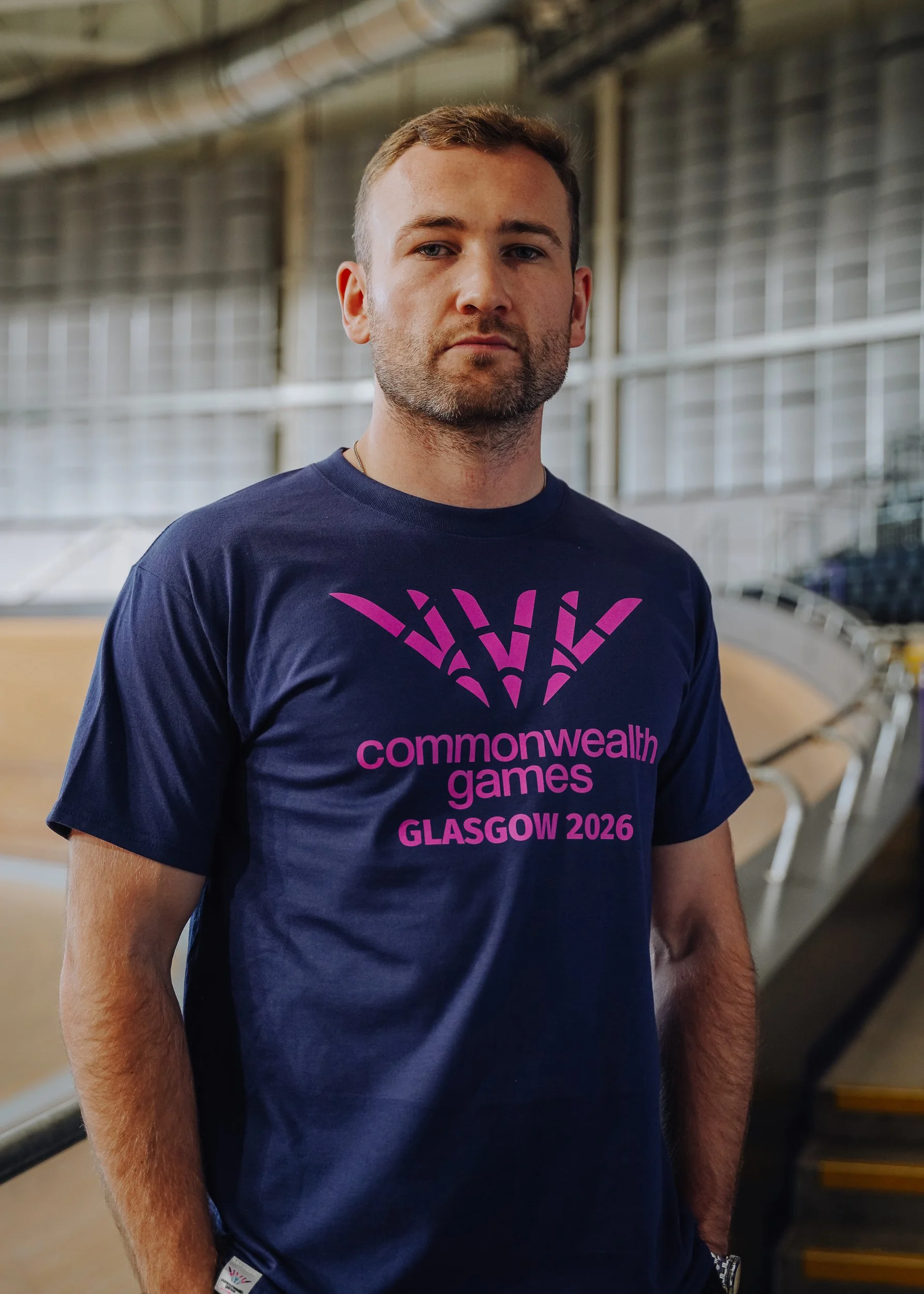 Man wearing navy blue t-shirt with pink logo and text for 2026 Glasgow Commonwealth Games inside an indoor sports arena.