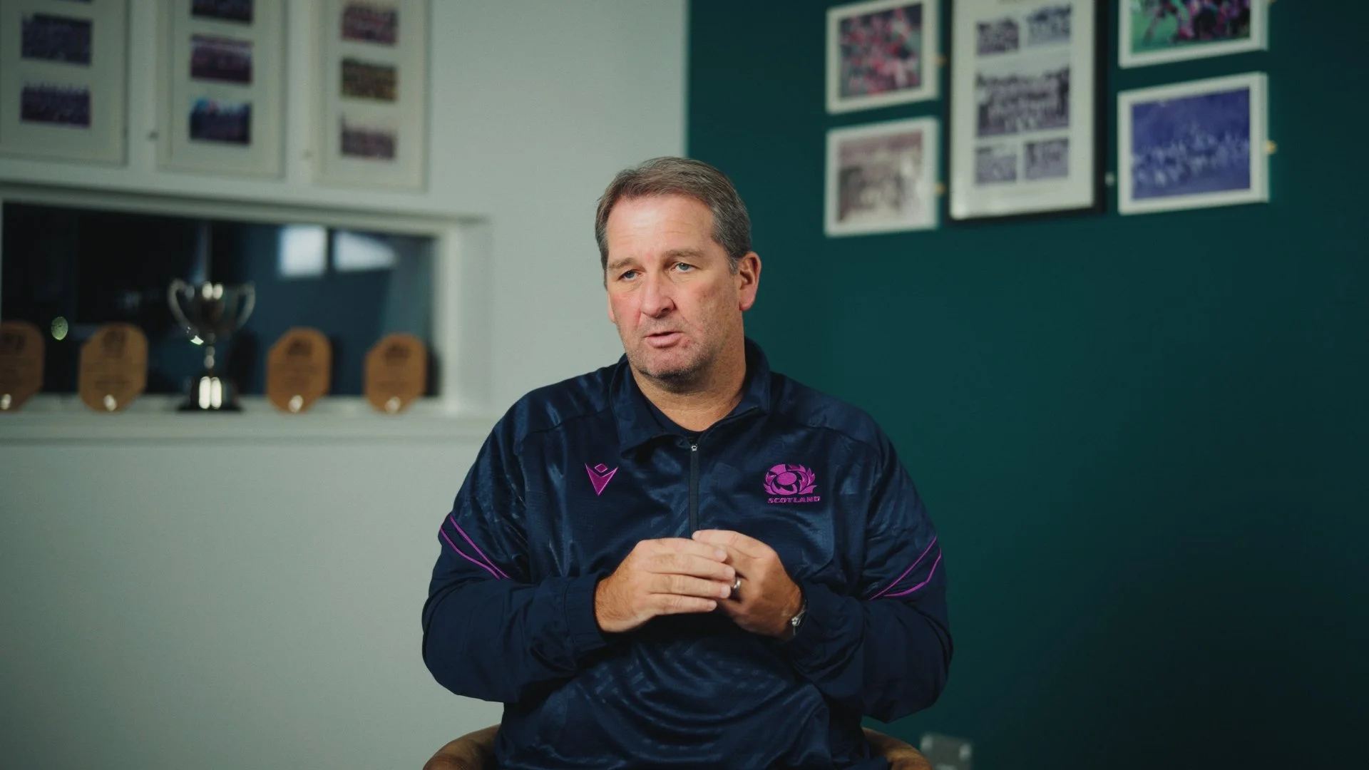 A man wearing a Scotland rugby jacket sitting in a room decorated with framed pictures and trophies on a shelf.