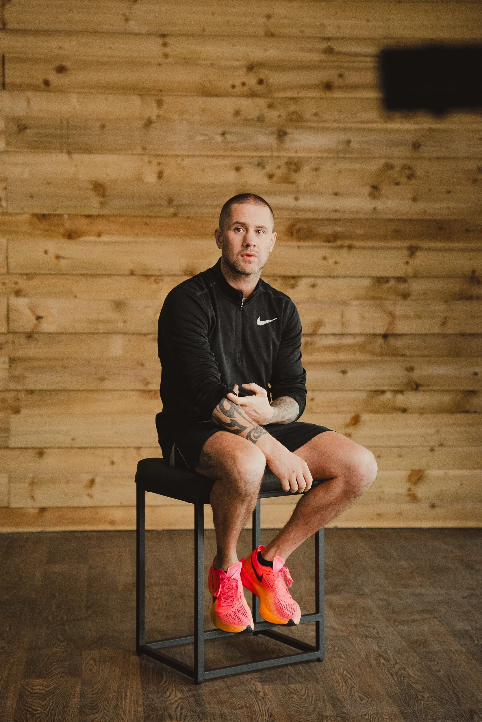 A man sitting on a black stool in front of a wooden wall, wearing a black Nike sports jacket, black shorts, and bright pink running shoes.