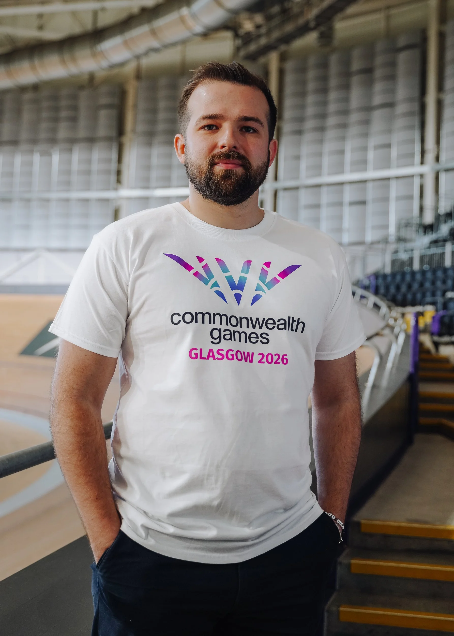 A man with a beard and short brown hair stands inside a sports arena, wearing a white t-shirt with the text 'Commonwealth Games Glasgow 2026' and a colorful logo.