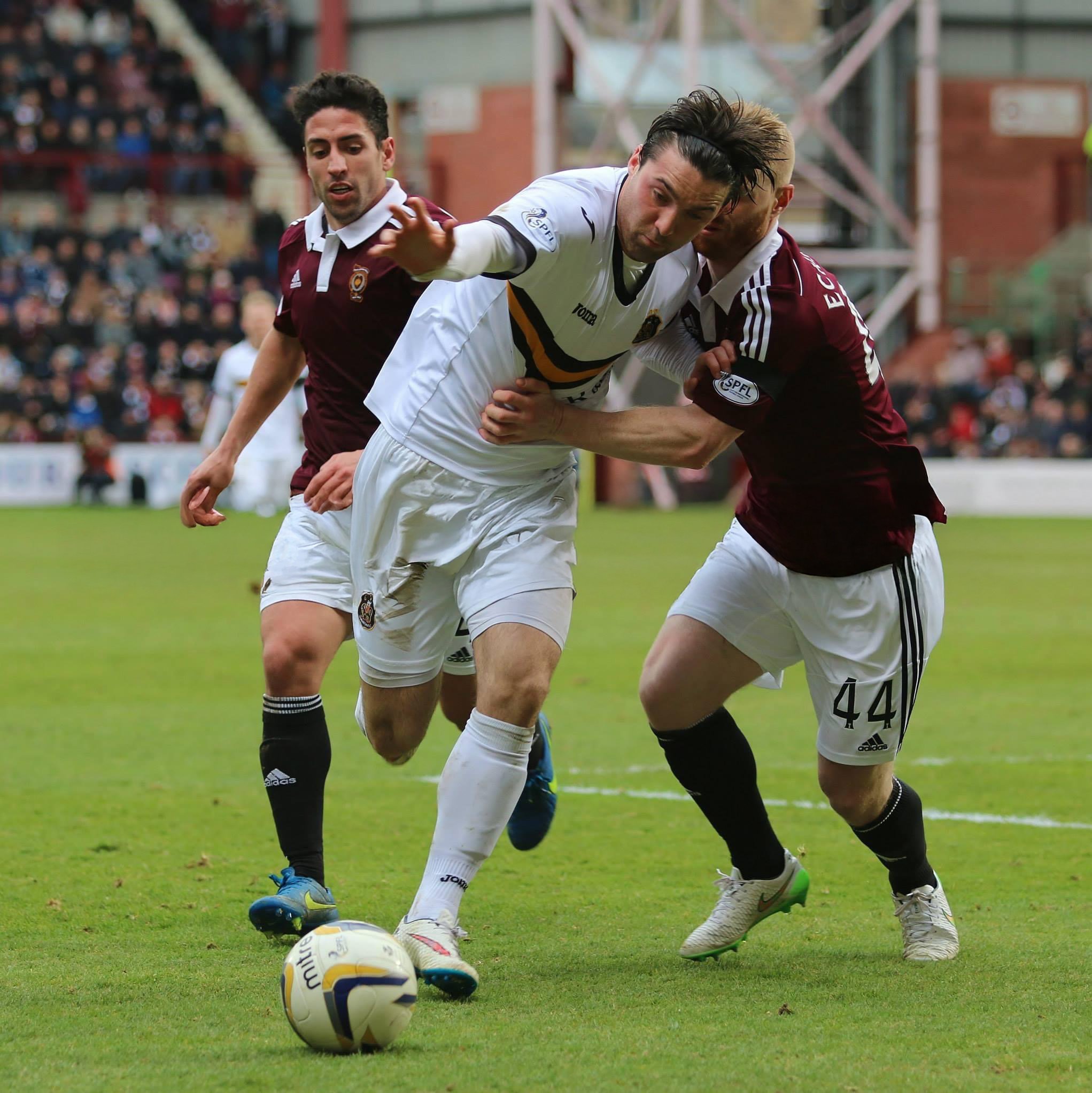 Two soccer players in maroon uniforms and one player in a white uniform are competing for possession of the soccer ball on a green field during a match, with spectators in the background.