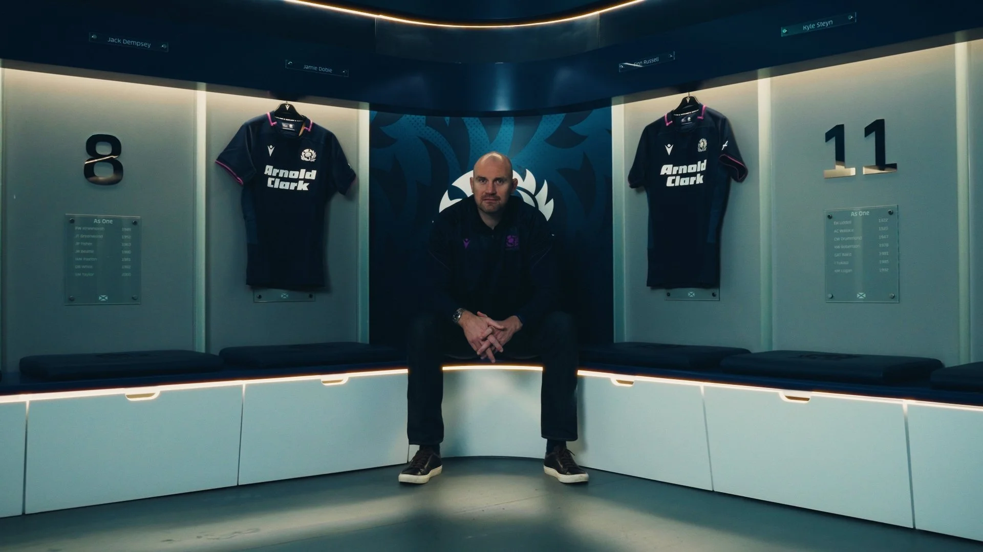 A man sitting in a sports locker room with two navy blue rugby jerseys hanging behind him. The jerseys feature the Arnold Clark logo. The lockers are numbered 8 and 11 and have plaques listing famous players and their achievements.