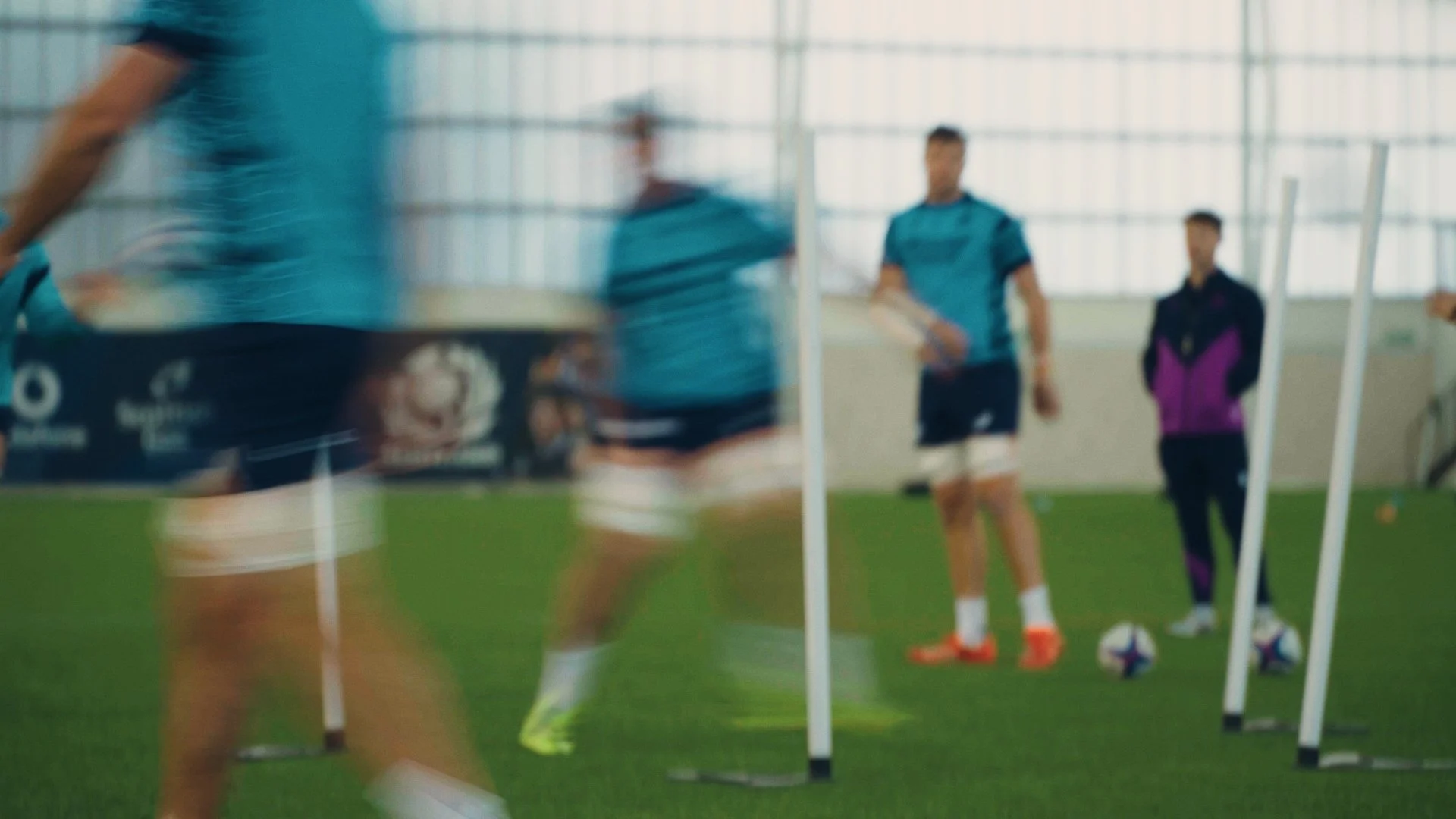 Soccer players training indoors with agility poles, with one player in focus and others blurred in background.