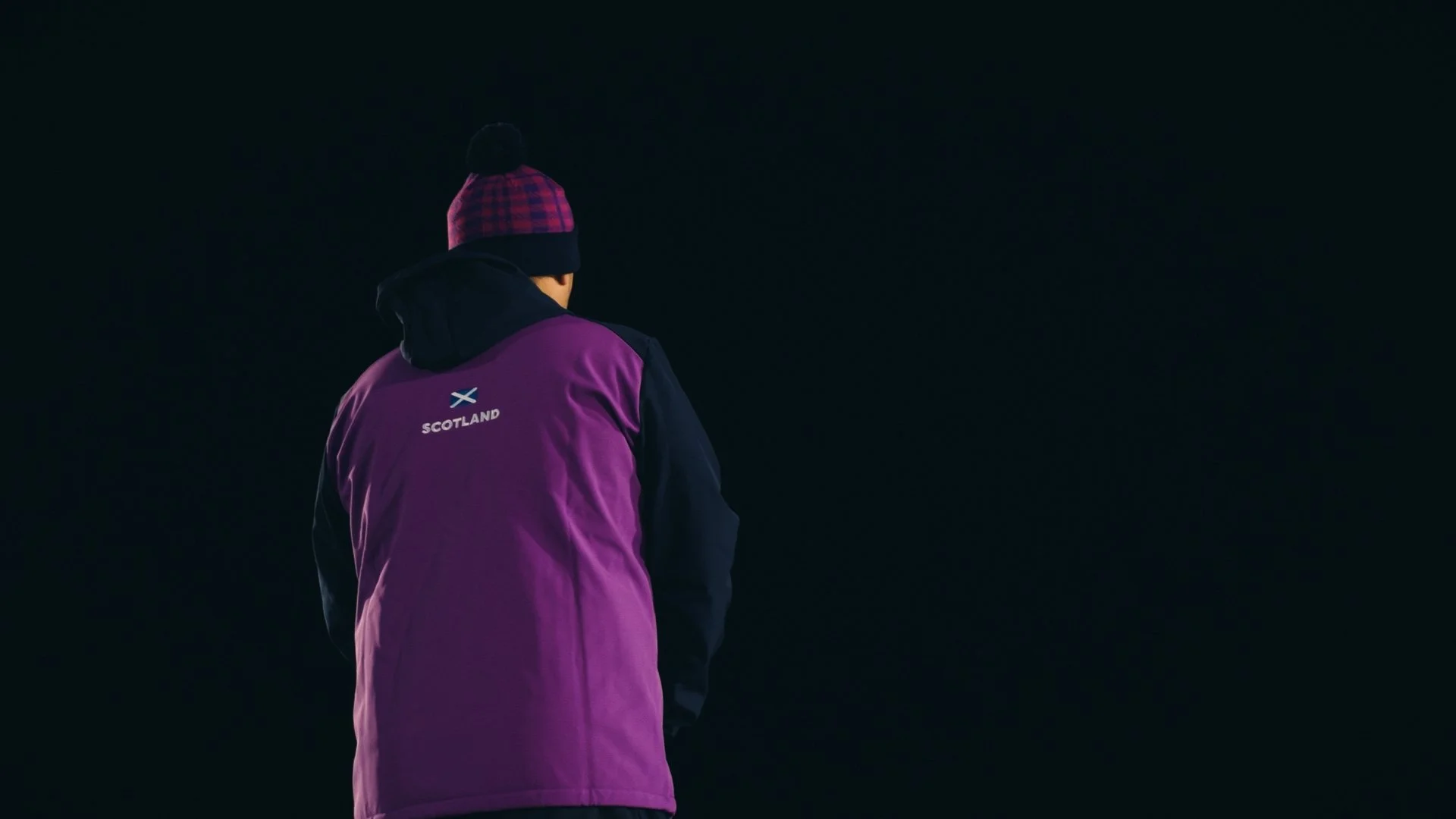 Person wearing a purple jacket with 'Scotland' and the Scottish flag on the back, standing outdoors at night.