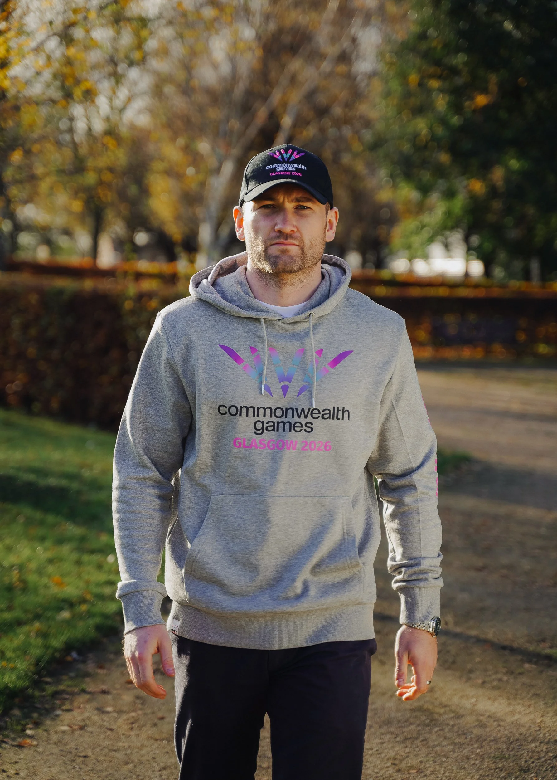 A man wearing a gray hoodie and a black cap, walking outdoors on a dirt path, with trees and fall foliage in the background. The hoodie and cap display the Commonwealth Games Glasgow 2026 logo.