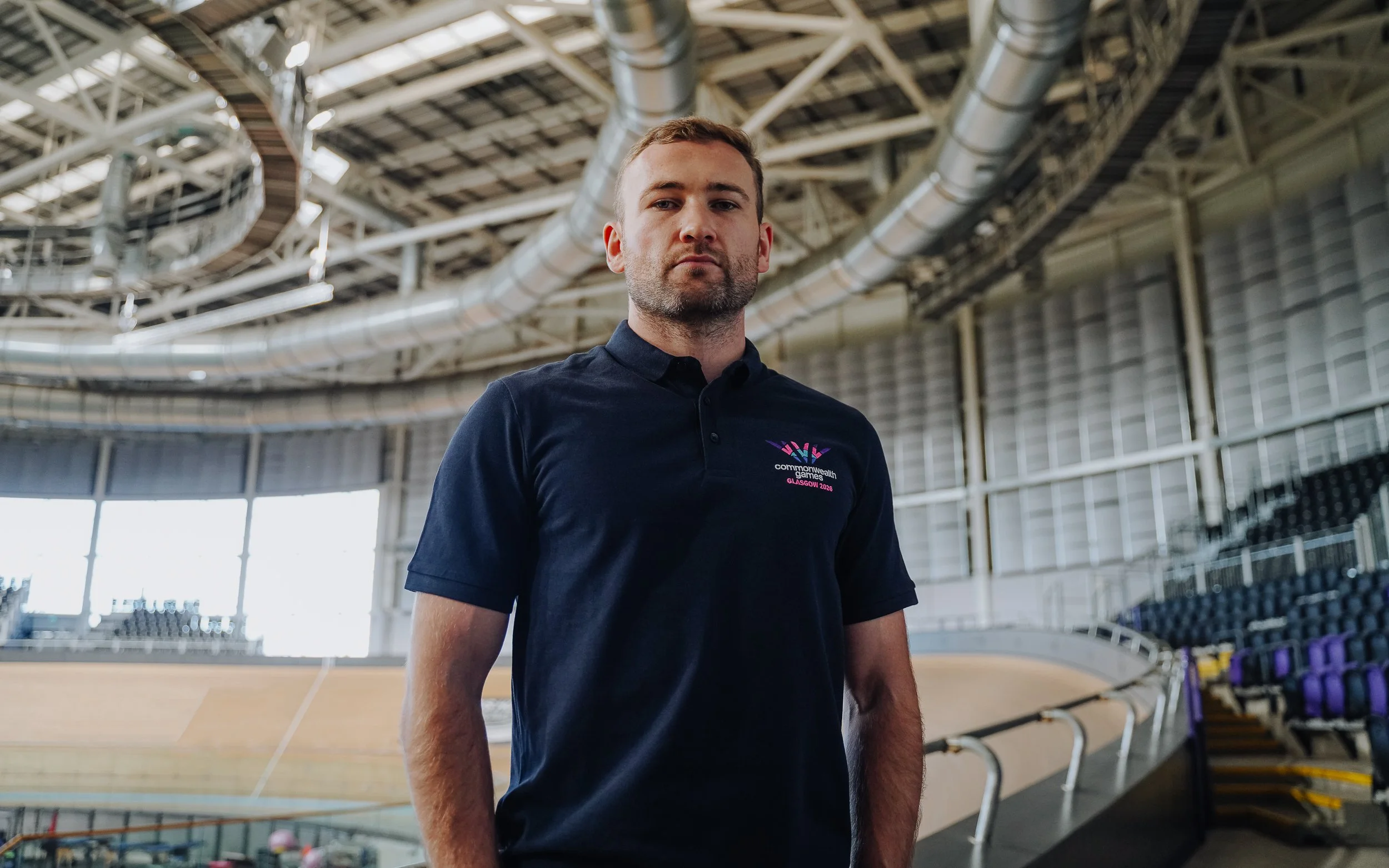 A man in a dark polo shirt standing inside an indoor velodrome with a curved wooden cycling track and seating in the background.