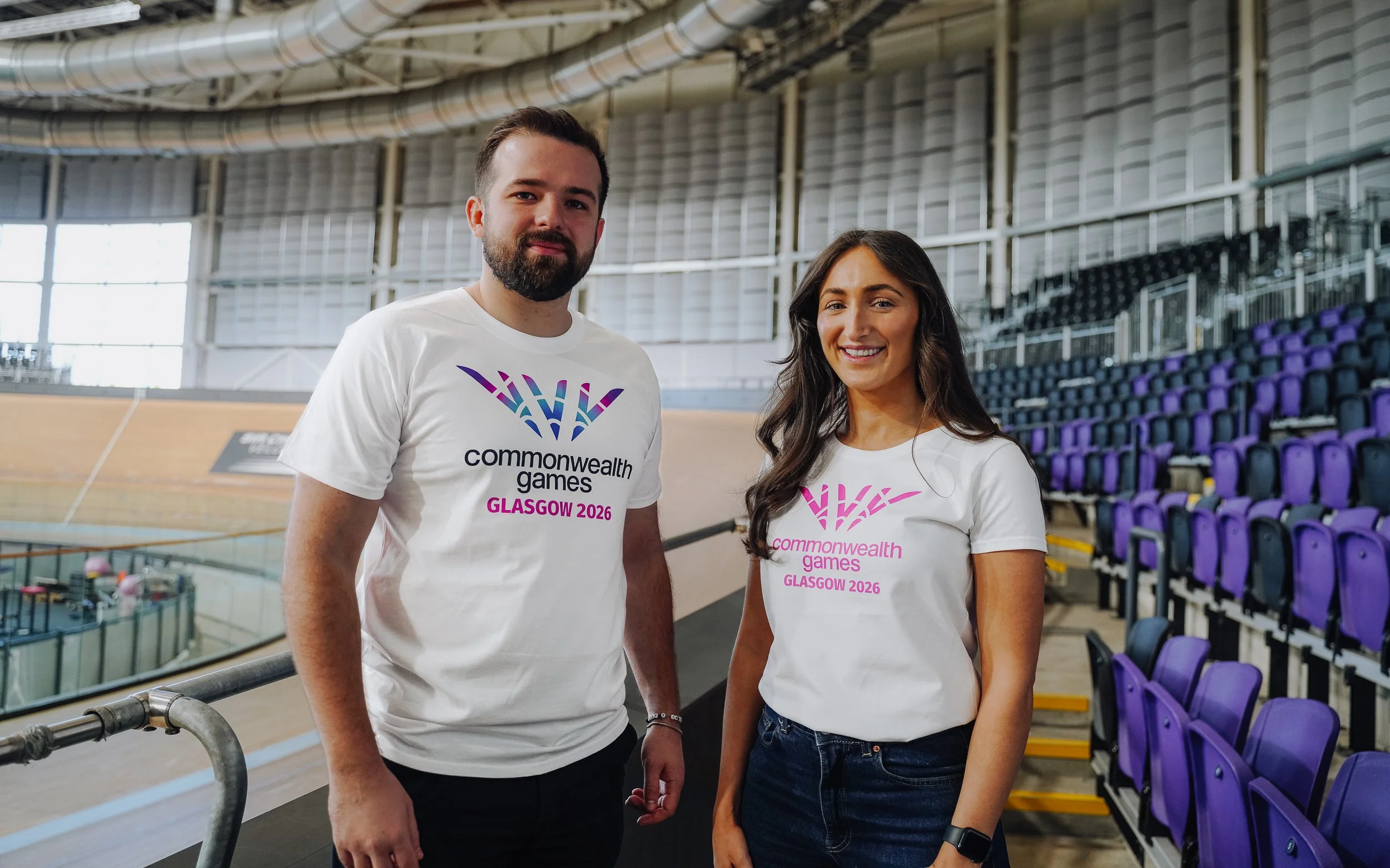 Two smiling individuals standing in an indoor velodrome, wearing white t-shirts with the Commonwealth Games Glasgow 2026 logo and text.