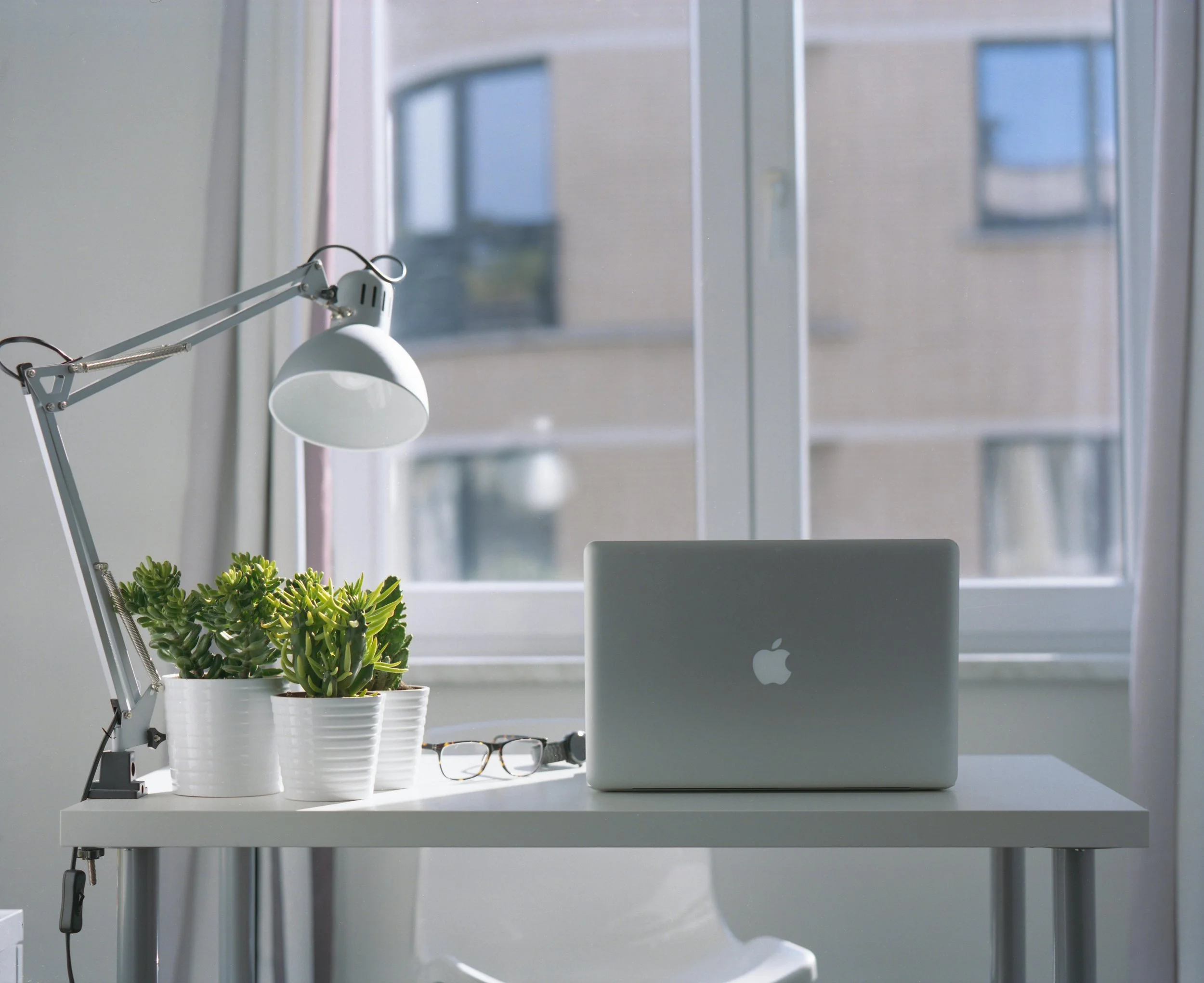 A minimalist workspace with a silver MacBook laptop on a white desk, two white pots with green succulents, a pair of glasses, a black watch, a white adjustable desk lamp, and a window showing an urban building outside.