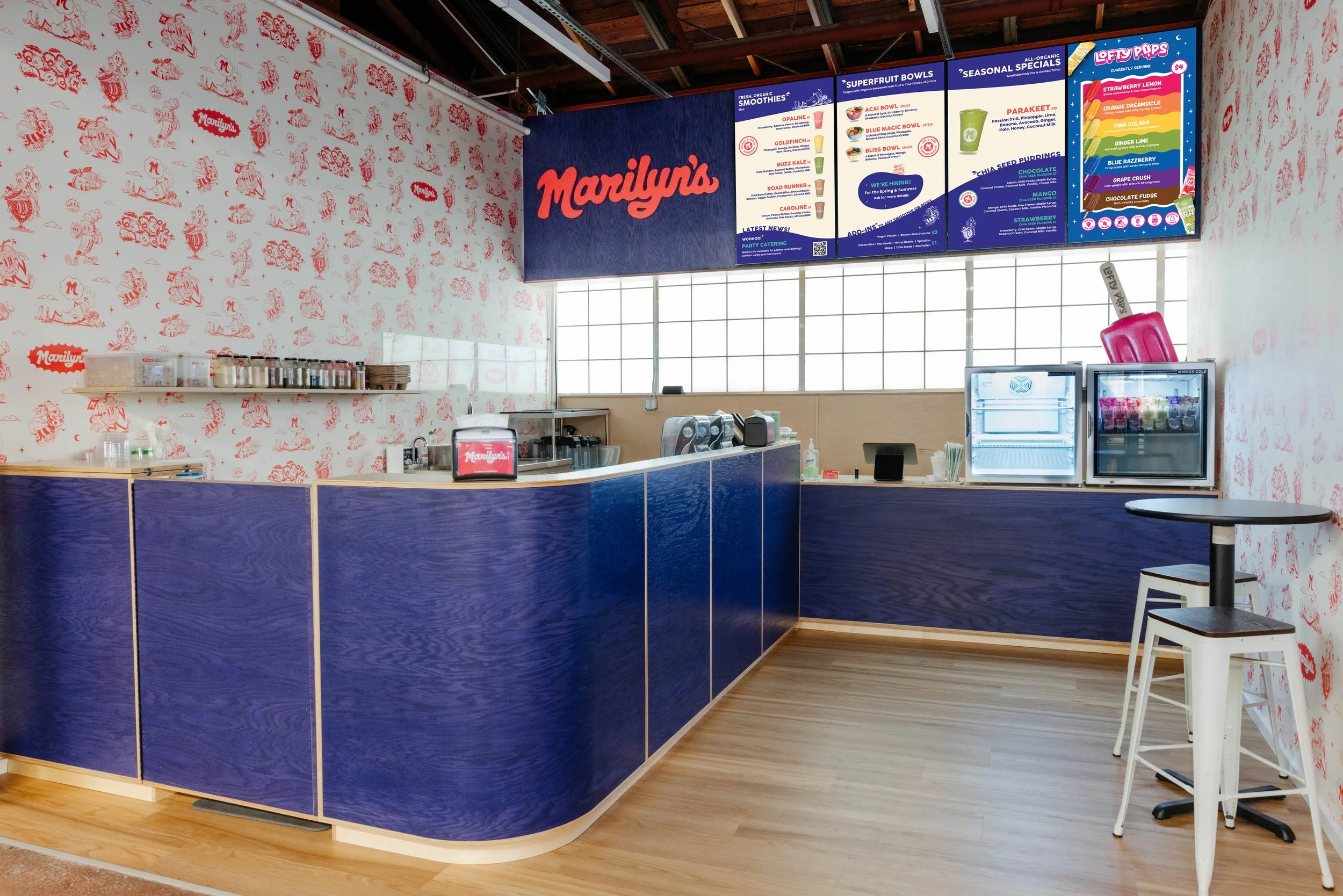 Smoothie and superfruit bowl shop interior with a menu board, pineapples, bananas, and potted plants. The menu lists smoothies and bowls with organic and vegan options. Social media information is displayed on the wall.