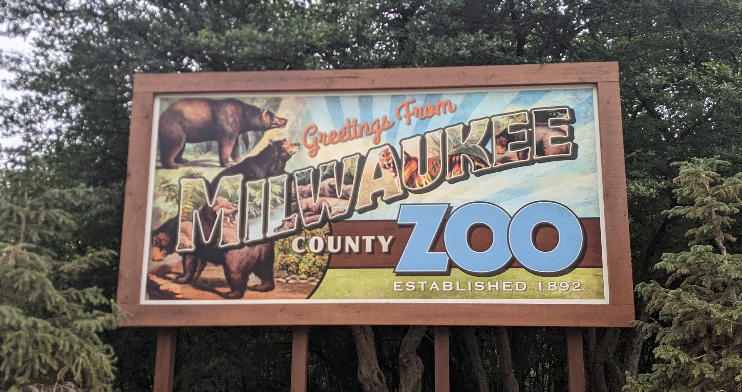 Signboard for Milwaukee County Zoo, established in 1892, featuring illustrations of bears and other animals, with trees and sky in the background.