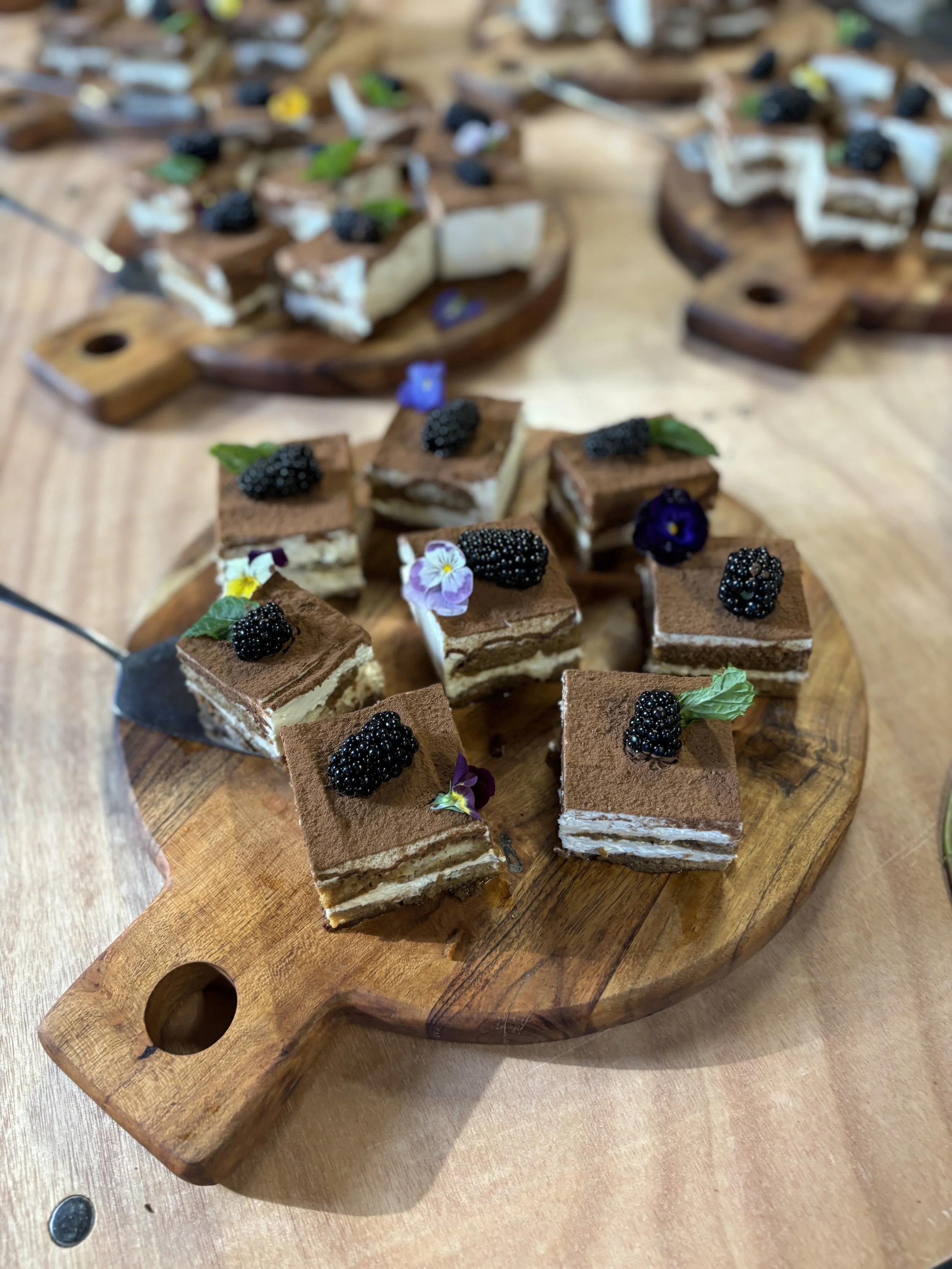 Dessert squares topped with blackberries and edible flowers on a wooden serving board.