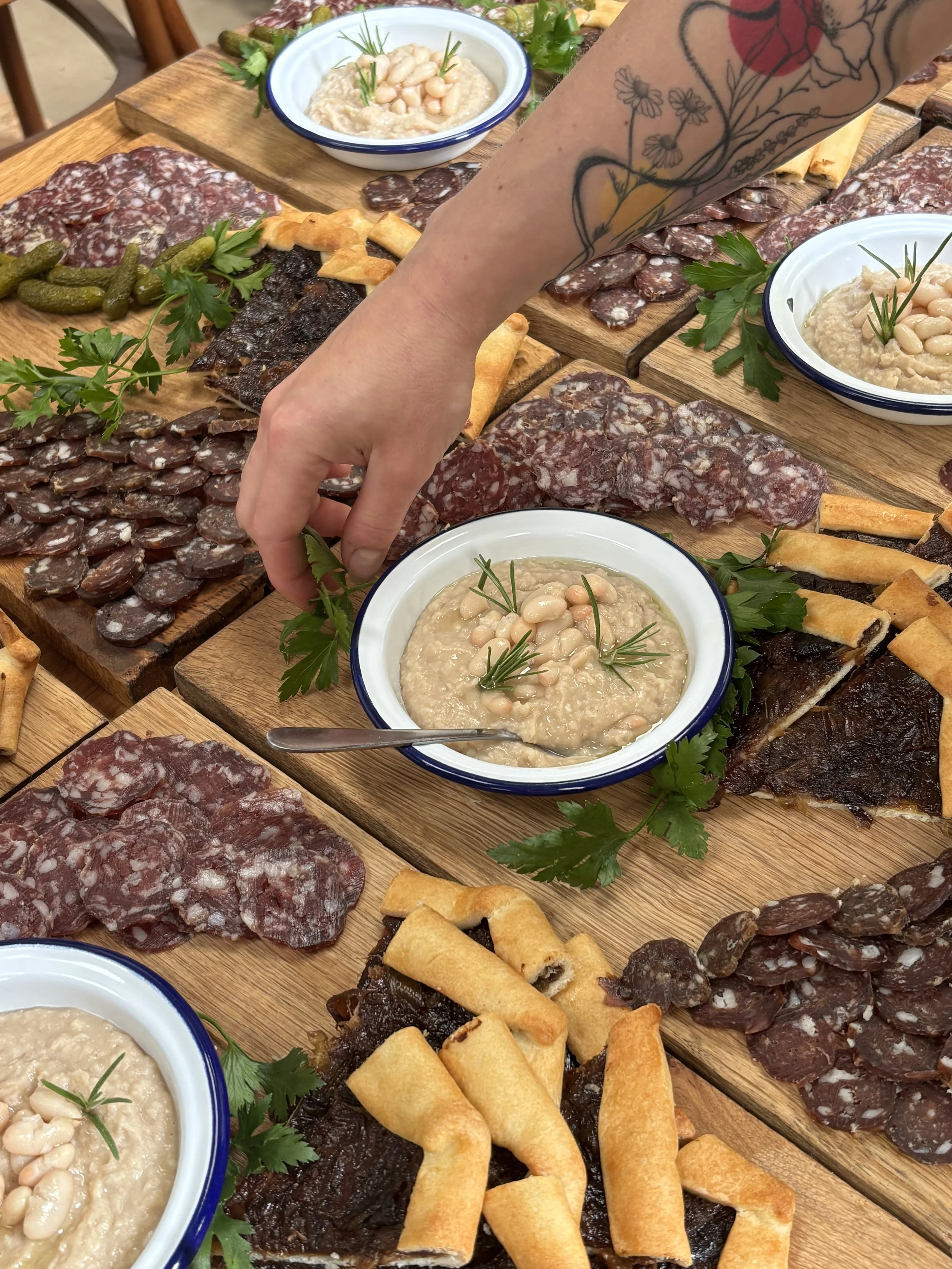 A wooden table with a charcuterie spread including various cured meats, peppered salami, pickles, cheese-filled pastry twists, and bowls of bean dip garnished with herbs.