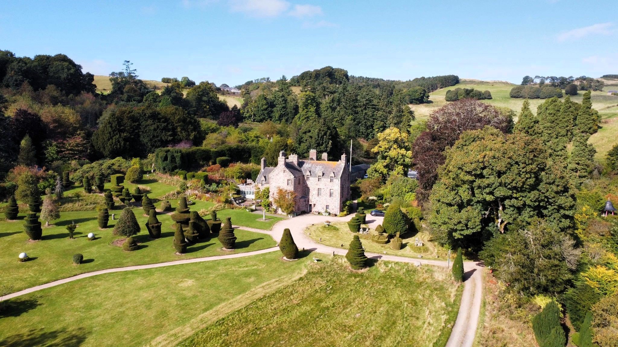 A large, historic stone mansion surrounded by a manicured garden with shaped trees and walking paths, set against a backdrop of rolling green hills and scattered trees under a bright blue sky.