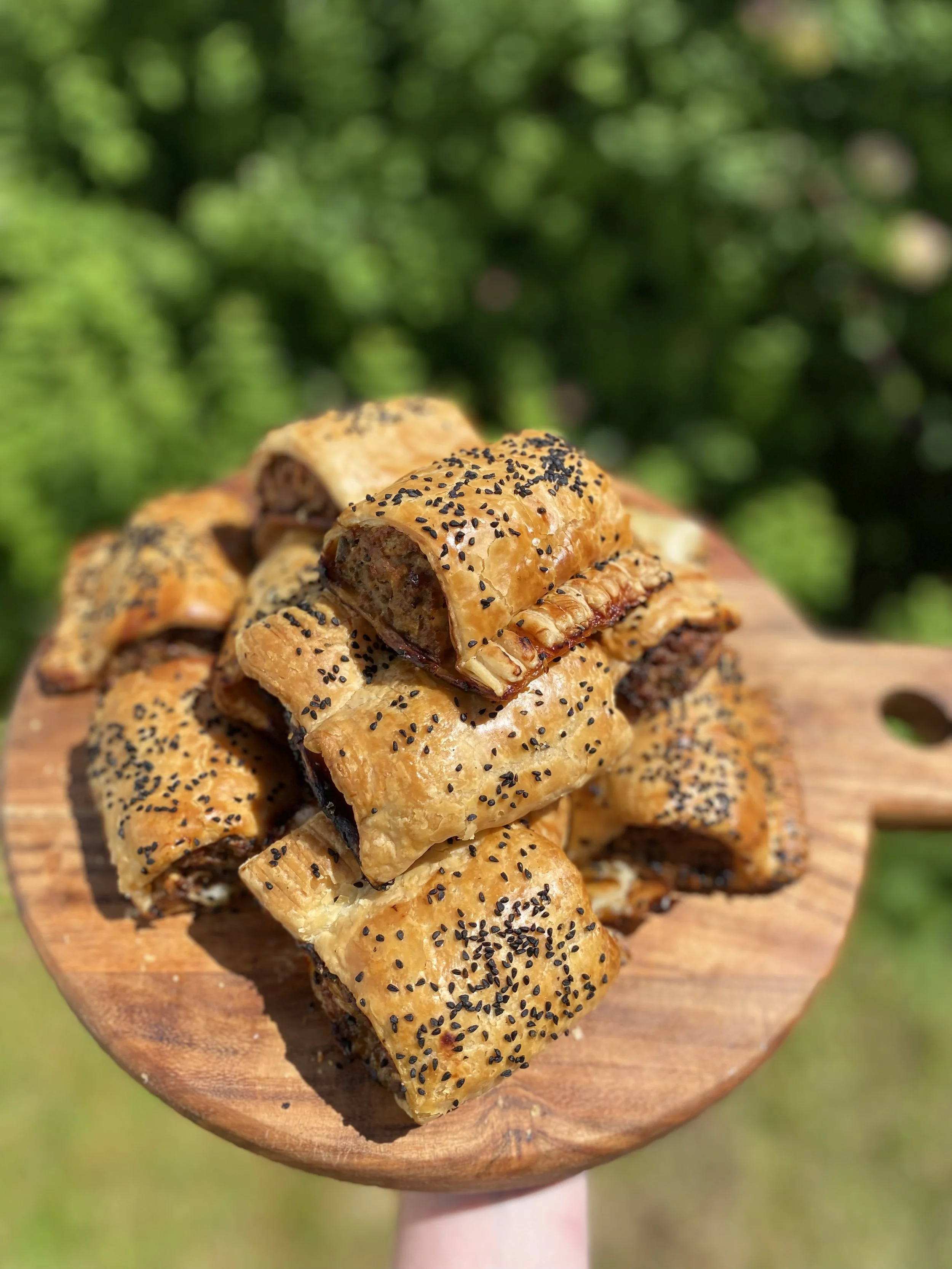 Close-up of sausage rolls with golden brown pastry and black sesame seeds on top, stacked on a wooden serving board outdoors.