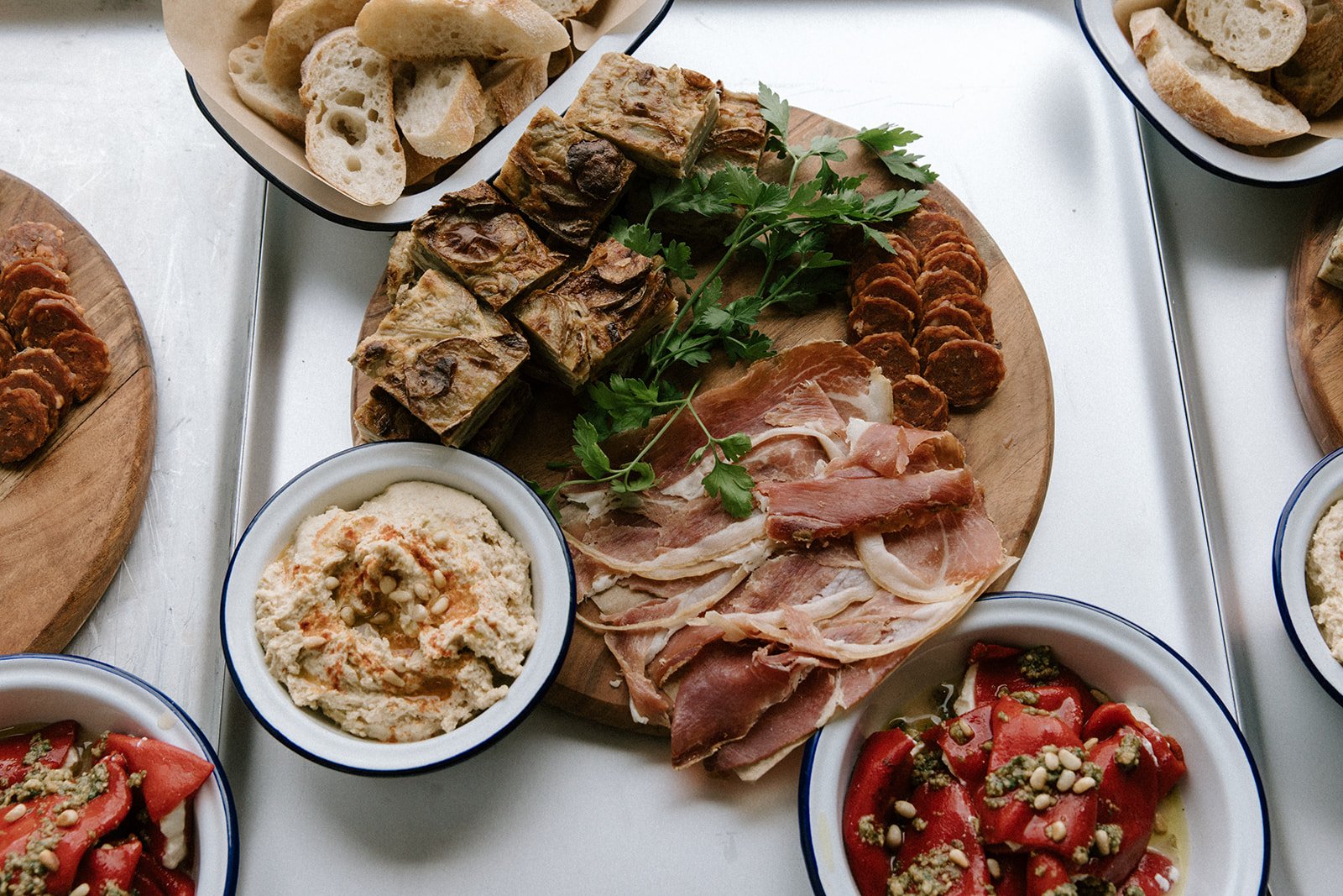 A charcuterie board with sliced cured meats, bread, and herbs on a wooden platter; surrounded by bowls of hummus and tomato salad.