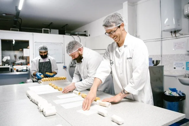 People baking cookies in a commercial kitchen, wearing hairnets and aprons, working on cookie dough.