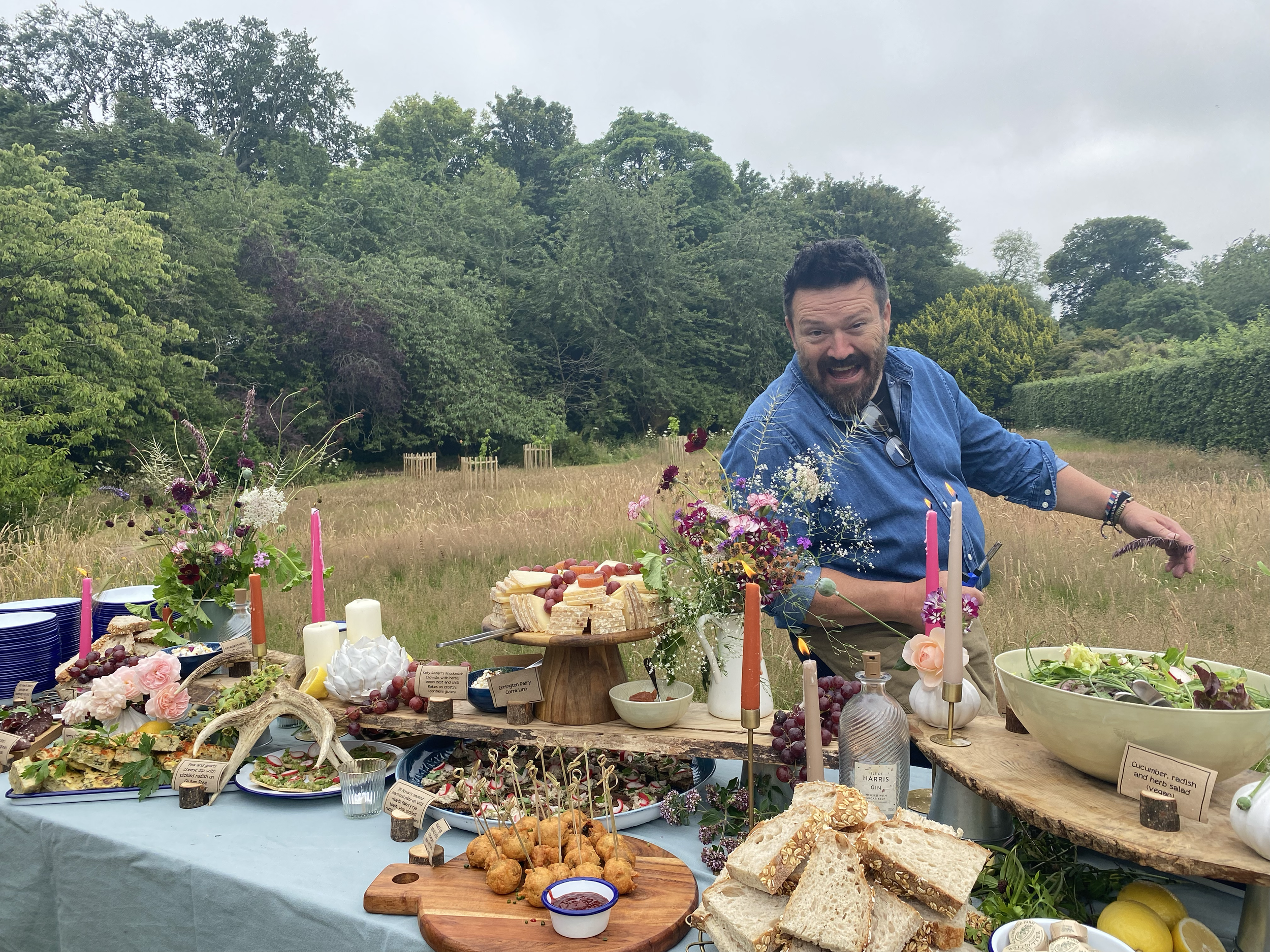 A man with dark hair and beard wearing a blue shirt enjoying a picnic outdoors. In front of him, there is a table filled with various foods including a cheese platter, bread, and salads, decorated with flowers and candles. The background features lus