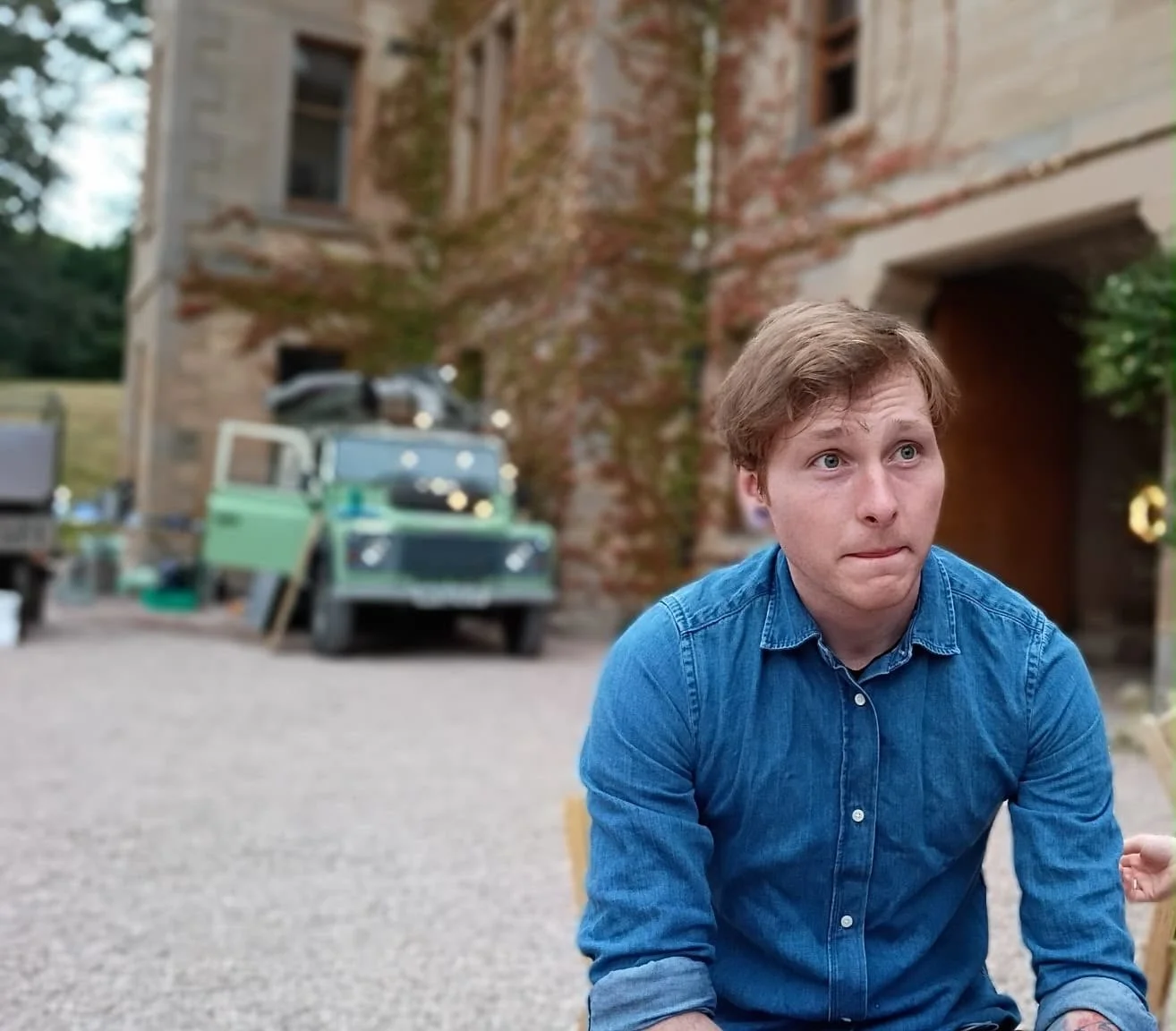 A young man with reddish-brown hair and a blue denim shirt is in the foreground, with a slightly surprised or contemplative expression, outdoors near a large brick building with arched entryways and some vintage vehicles in the background.