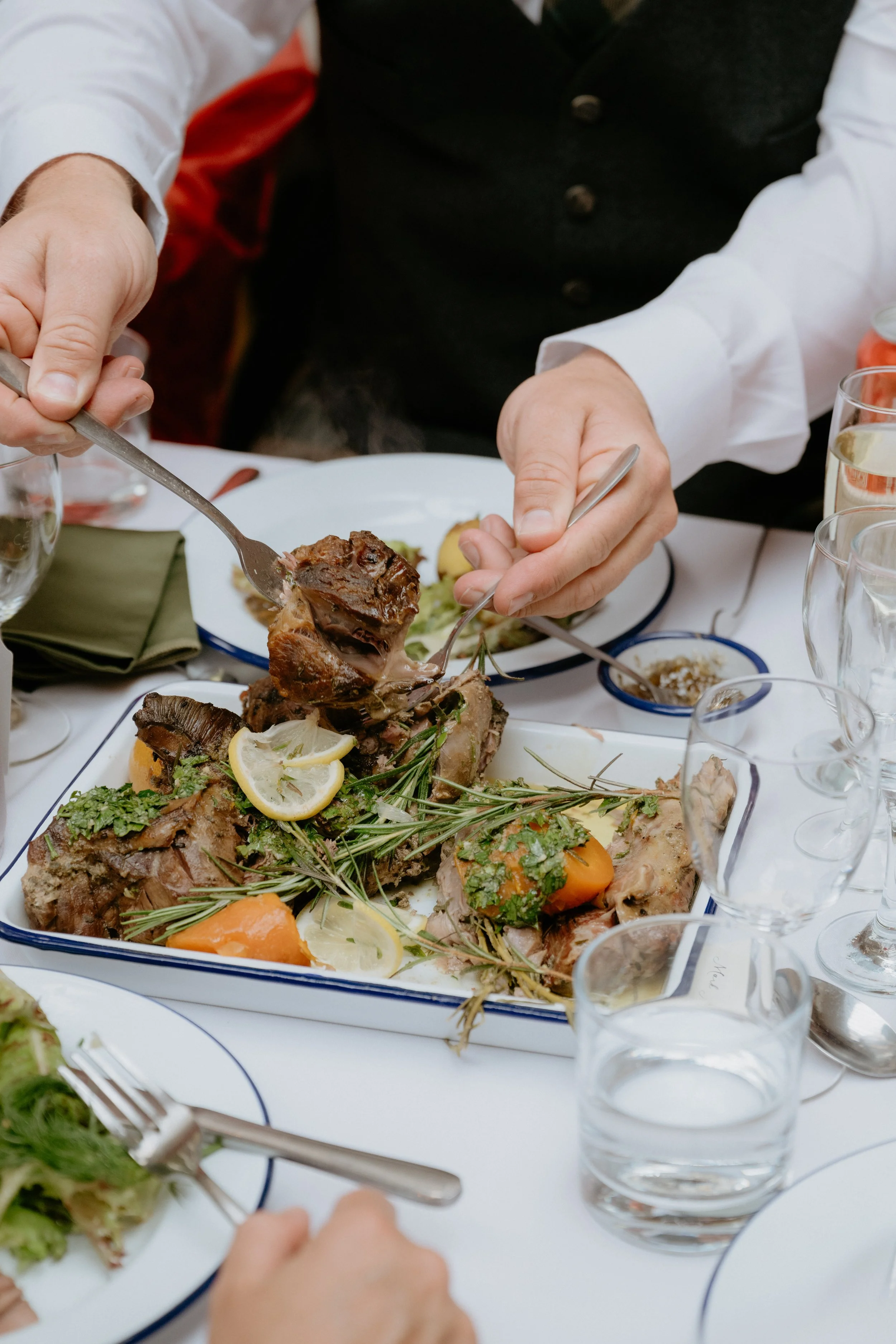Hands serving carved meat and vegetables from a platter at a dinner table with glasses of water and a green napkin nearby.
