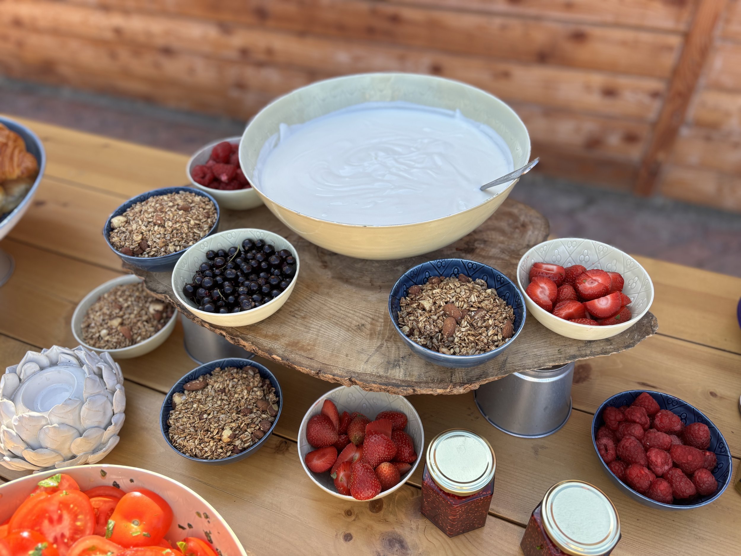 Various bowls containing yogurt, berries, oats, granola, and sliced strawberries on a wooden table, with a wooden wall in the background.