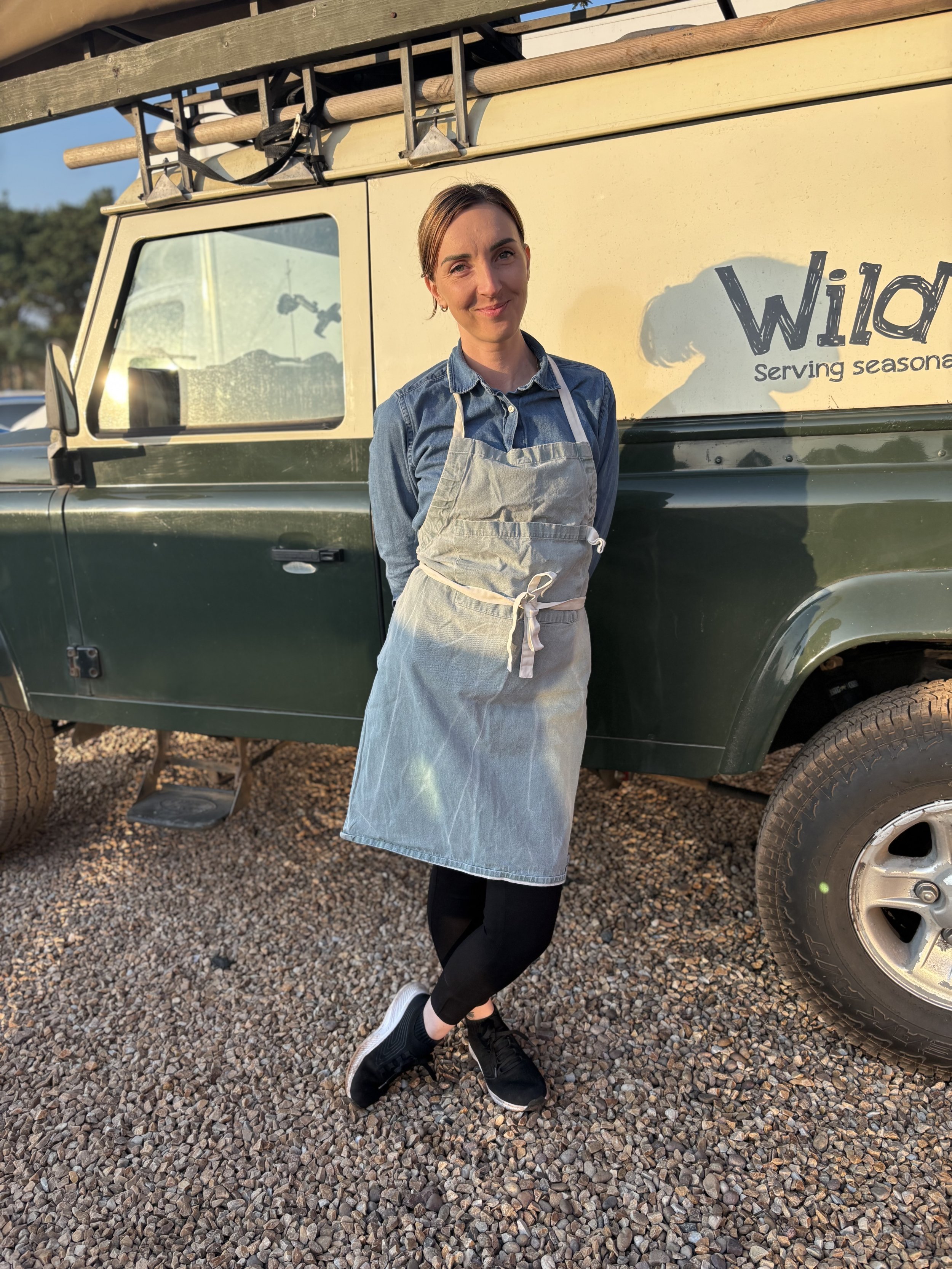 A woman standing outdoors next to a beige food truck with a sign that says 'Wild' and 'Serving season'. She is wearing a denim shirt, a gray apron, black pants, and sneakers, with her hands behind her back and a slight smile.