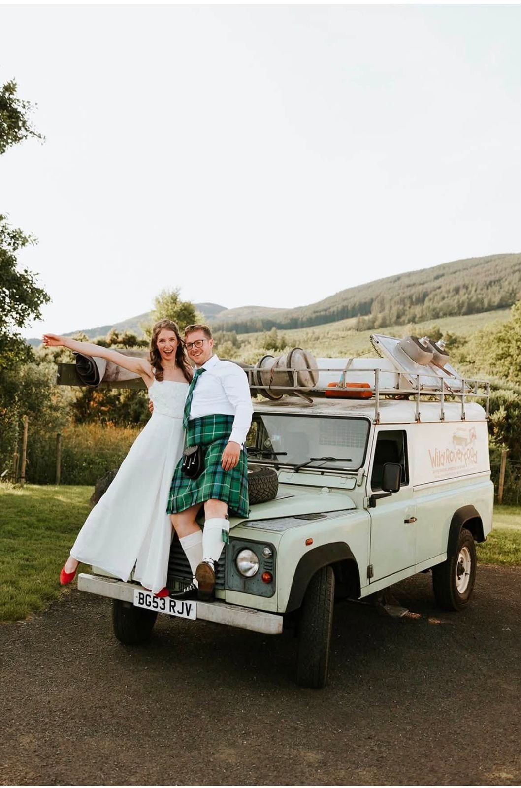 A couple dressed in wedding attire standing on the front of a vintage light green utility vehicle, smiling and embracing outdoors with rolling hills and trees in the background.