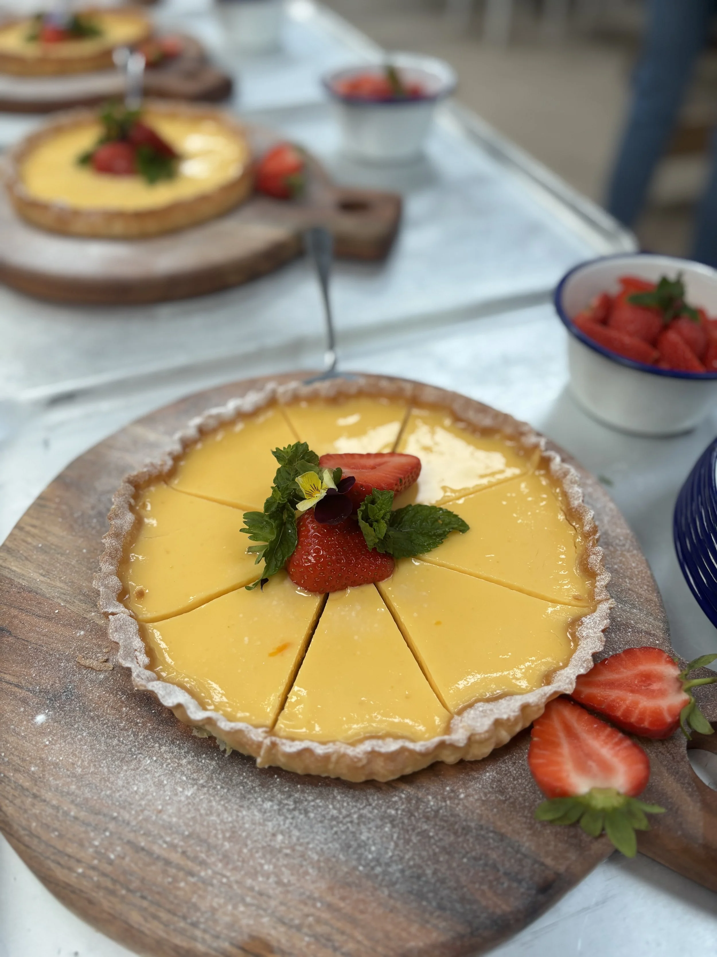 A yellow lemon tart garnished with strawberry slices and mint leaves on a wooden serving board, with strawberries and bowls in the background.